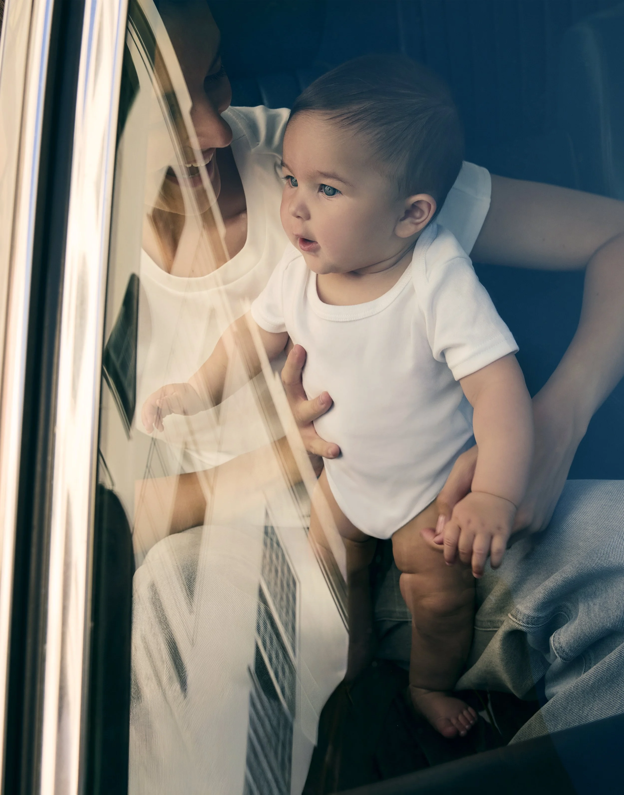A young boy with blue eyes and dark hair, wearing a white shirt, looks through a glass window on a train, with reflections and perspective.