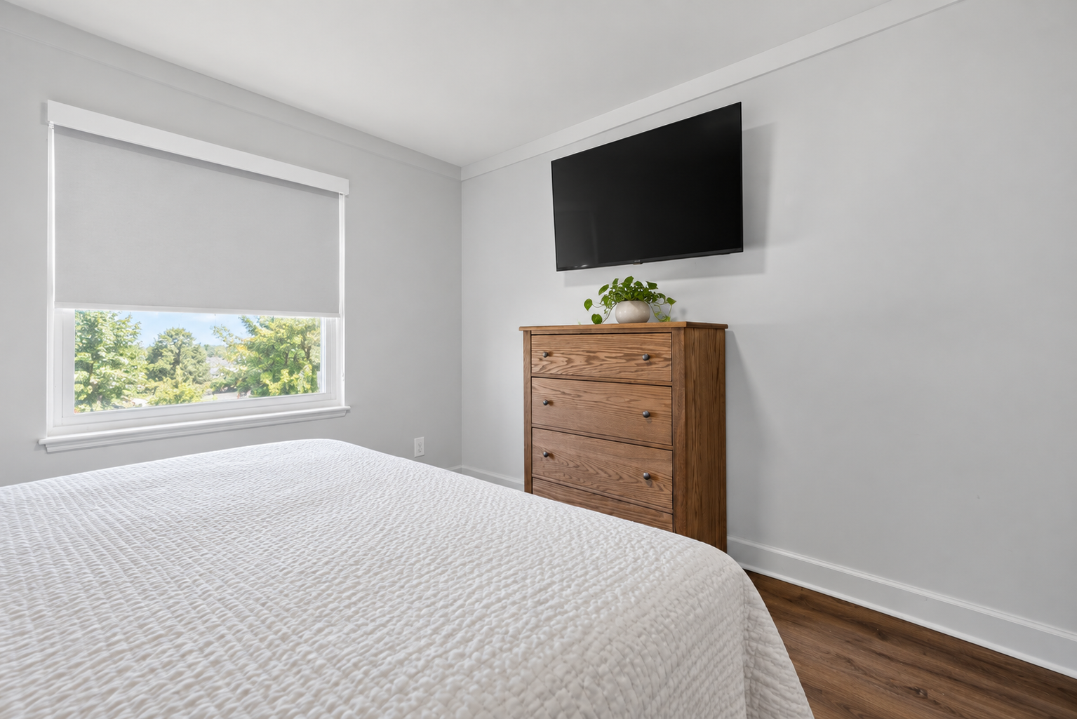 Bedroom at The Welcome Inn with a queen bed white bedspread, a window with a white shade, a wooden dresser, a potted plant, and a wall-mounted television.