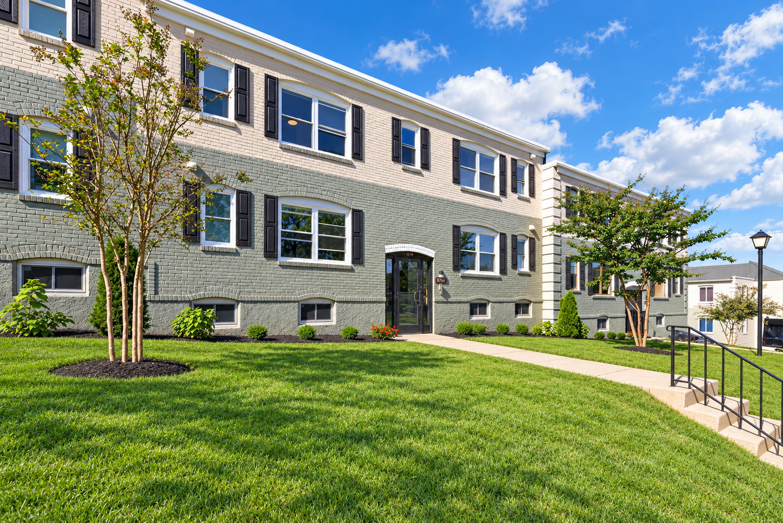 The Welcome Inn exterior with brick and siding exterior, multiple windows with black shutters, and a black front door. The building is surrounded by a well-maintained green lawn with trees, shrubs, and a concrete walkway with a metal handrail.