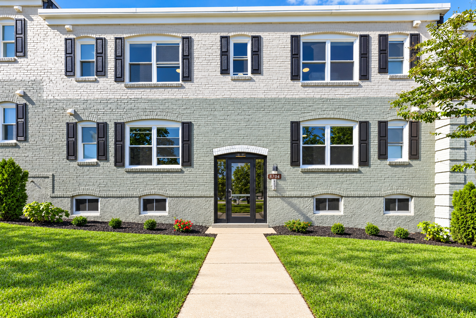 Front view of The Welcome Inn, a two-story extended stay hotel white brick exterior, black window shutters, and a black front door. Well-maintained green lawn and garden with small bushes and flowers. Clear blue sky and a tree on the right.