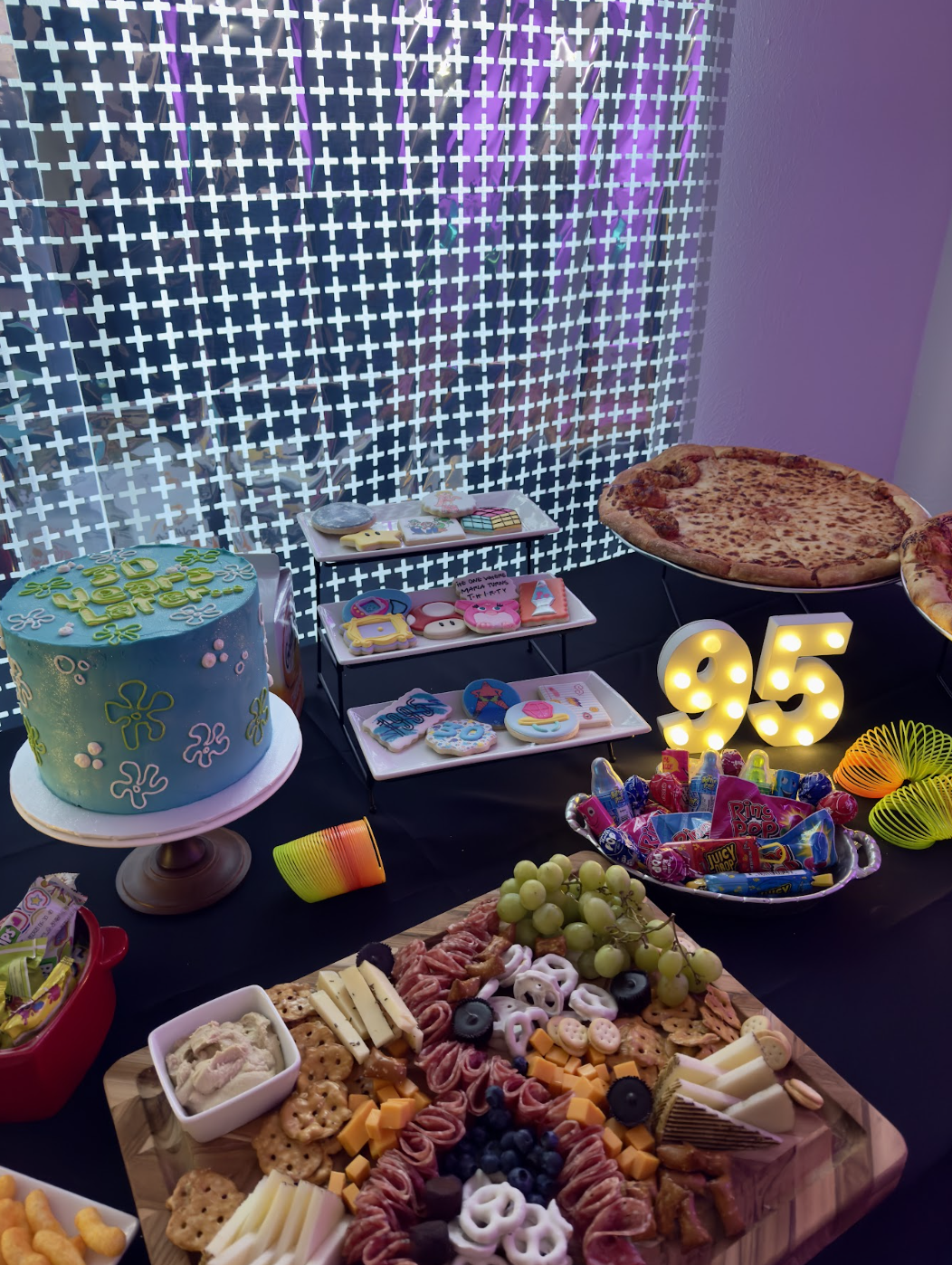 Birthday party setup with a cake, pizza, snacks, colorful candies, a cheese and meat platter, decorative numbers '95', and various treats on a table with a patterned backdrop.