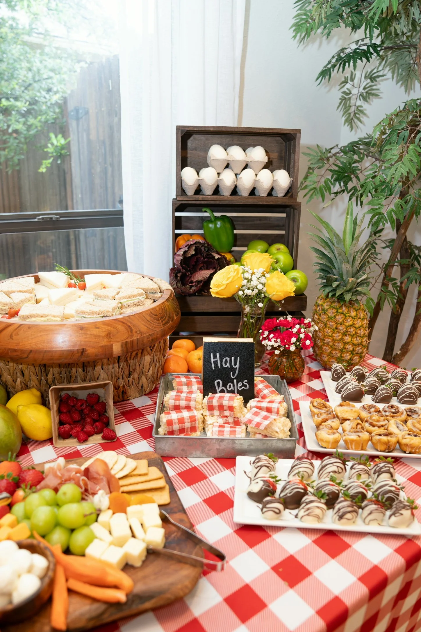 A table with a red and white checkered tablecloth displaying various desserts, fruits, and flowers, with a crate of eggs, pineapples, apples, strawberries, cheese, and pastries, along with a small chalkboard sign reading 'Hay Bales'.