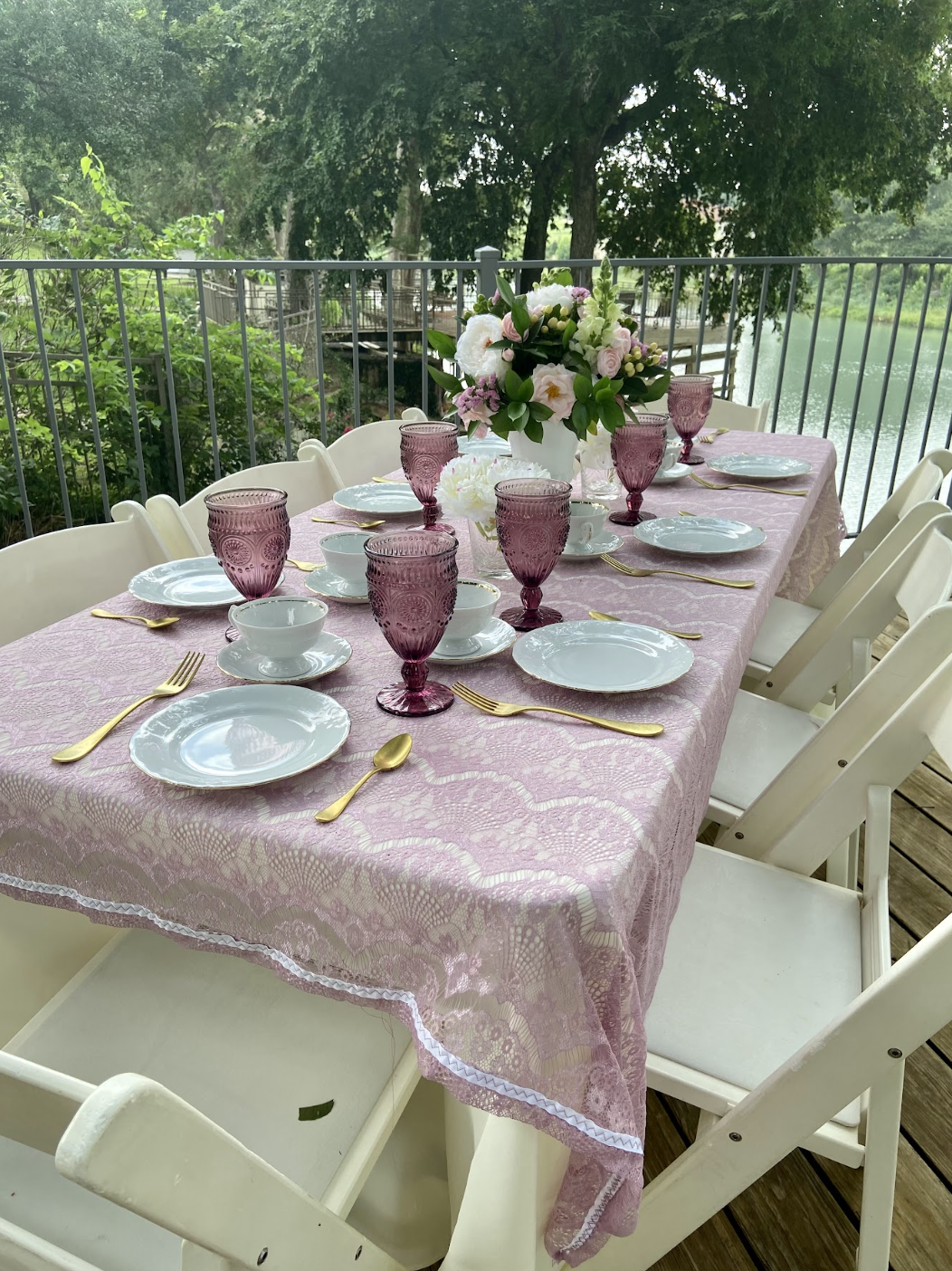 A decorated outdoor dining table with pink lace tablecloth, white plates, gold flatware, pink glass goblets, white bowls, and a large floral centerpiece on a deck overlooking a lake with trees.