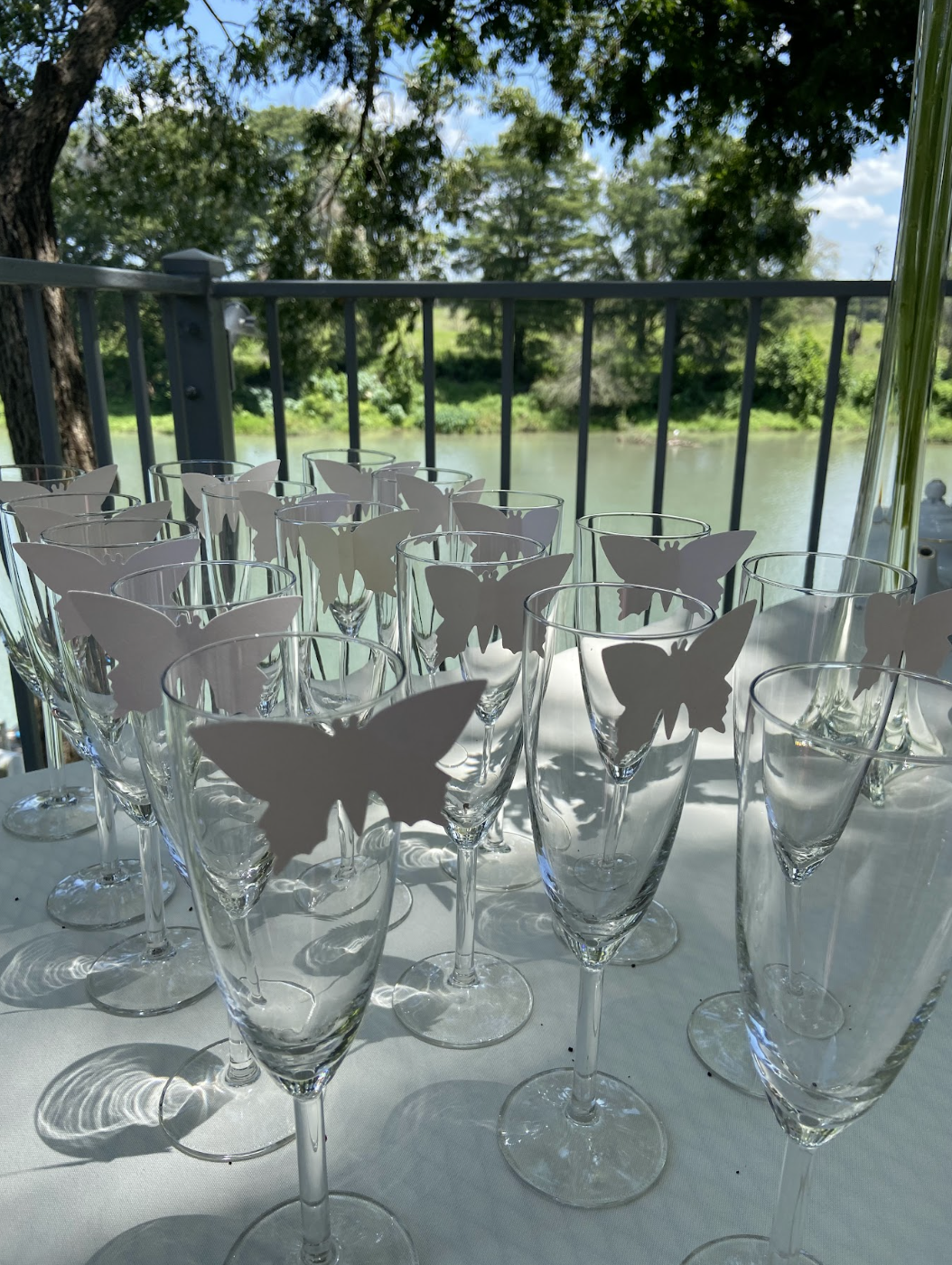 Multiple clear champagne flutes with paper butterfly decorations attached, set on a white tablecloth outdoors near a river and trees.
