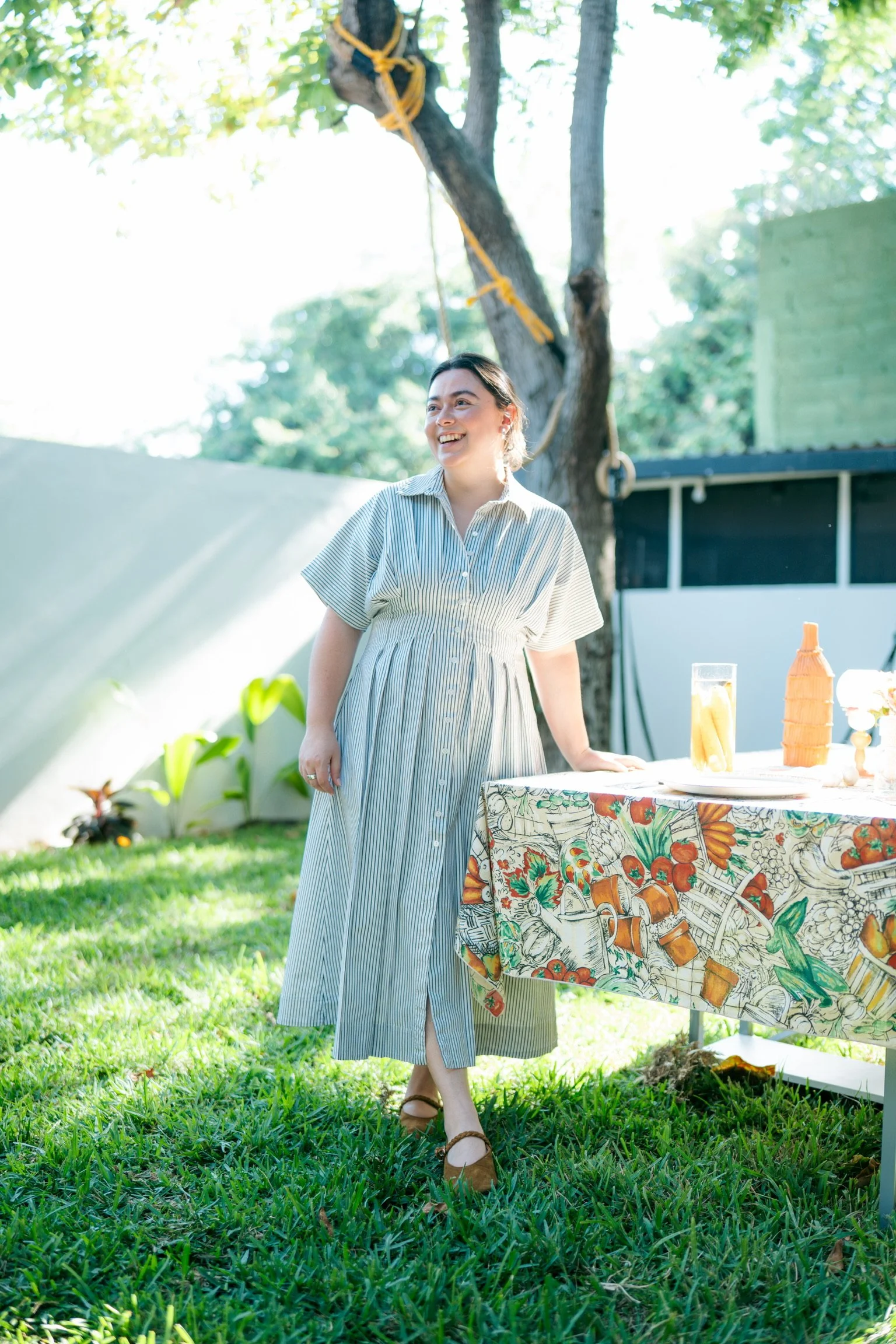 A woman in a long striped dress standing outdoors near a table with a colorful tablecloth. She is smiling and looking to her left. There is a large tree with yellow rope knots in the background, with sunlight filtering through the leaves.