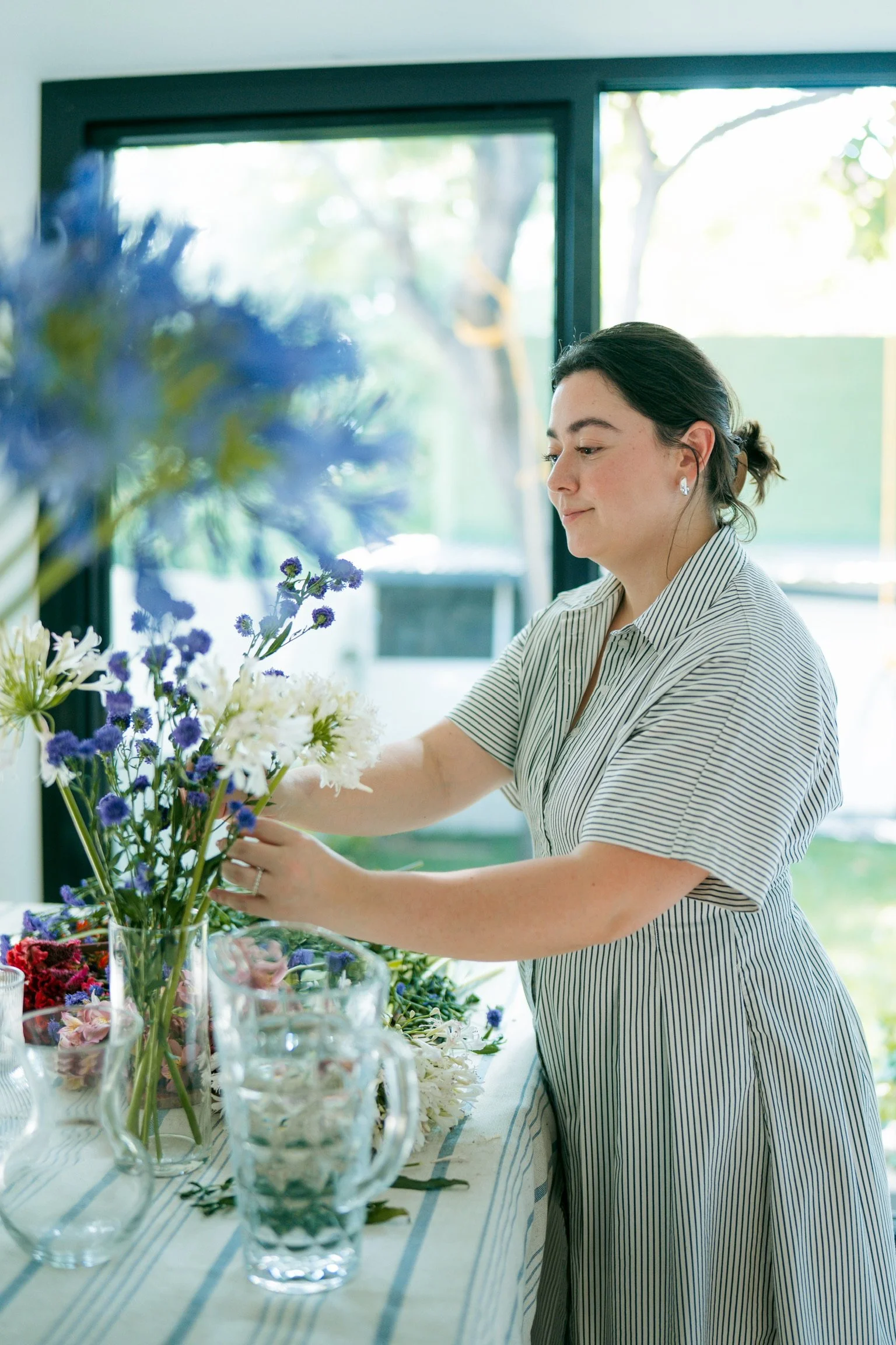 A woman arranging a bouquet of flowers at a table filled with various flower arrangements, near a large window with a view of trees outside.