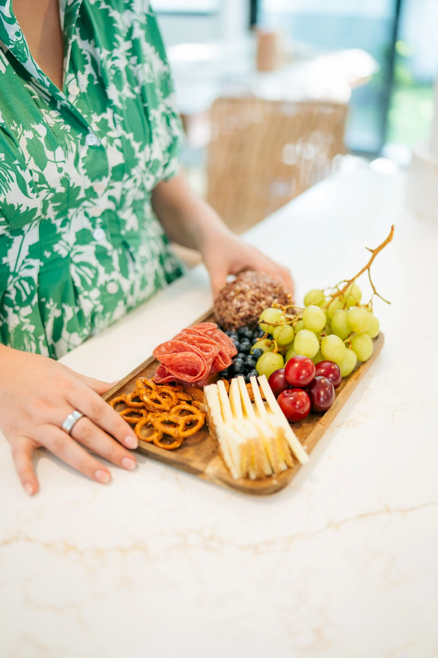 Person wearing a green and white patterned shirt arranging a wooden platter of assorted snacks, including grapes, cherry tomatoes, cheese sticks, pretzels, sliced meats, and a round snack ball, on a white kitchen counter, with a blurred background of a kitchen dining area.