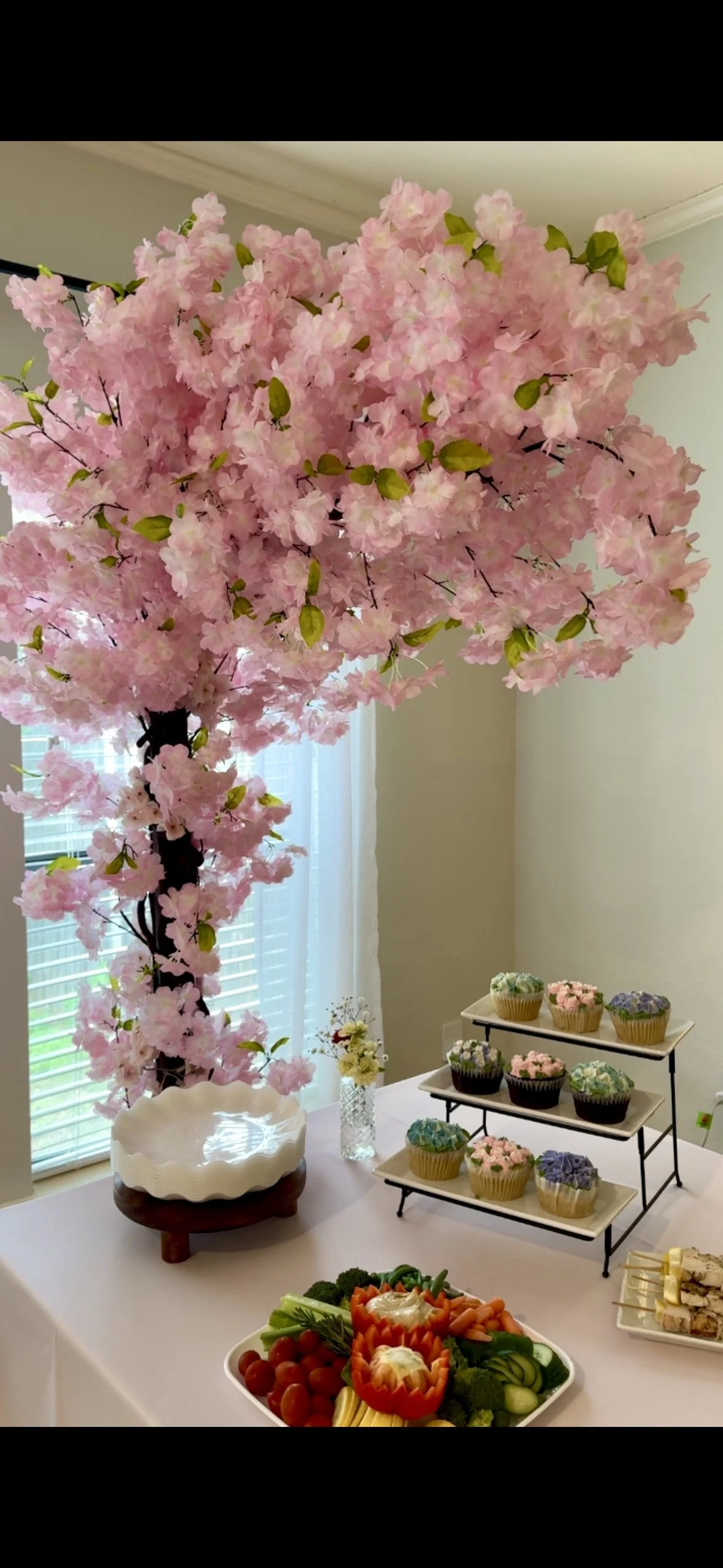 Decorative pink cherry blossom tree with artificial flowers at a party table, featuring cupcakes with pastel icing on a tiered stand, a tray of vegetables with creamy dip, and floral arrangements near a window.