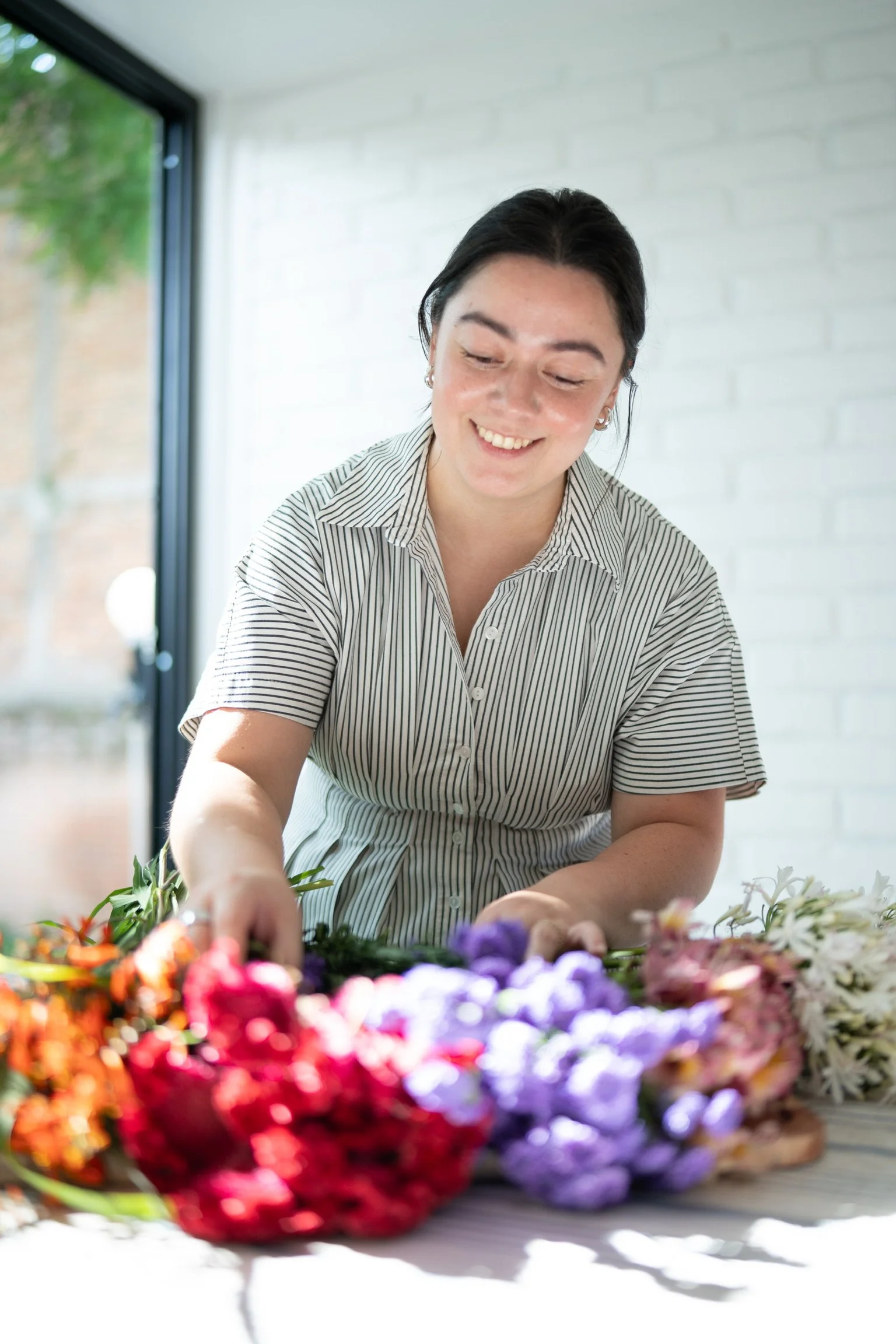 A woman smiling and arranging colorful flowers on a table in a bright room with a white brick wall and a large window showing green outdoors.