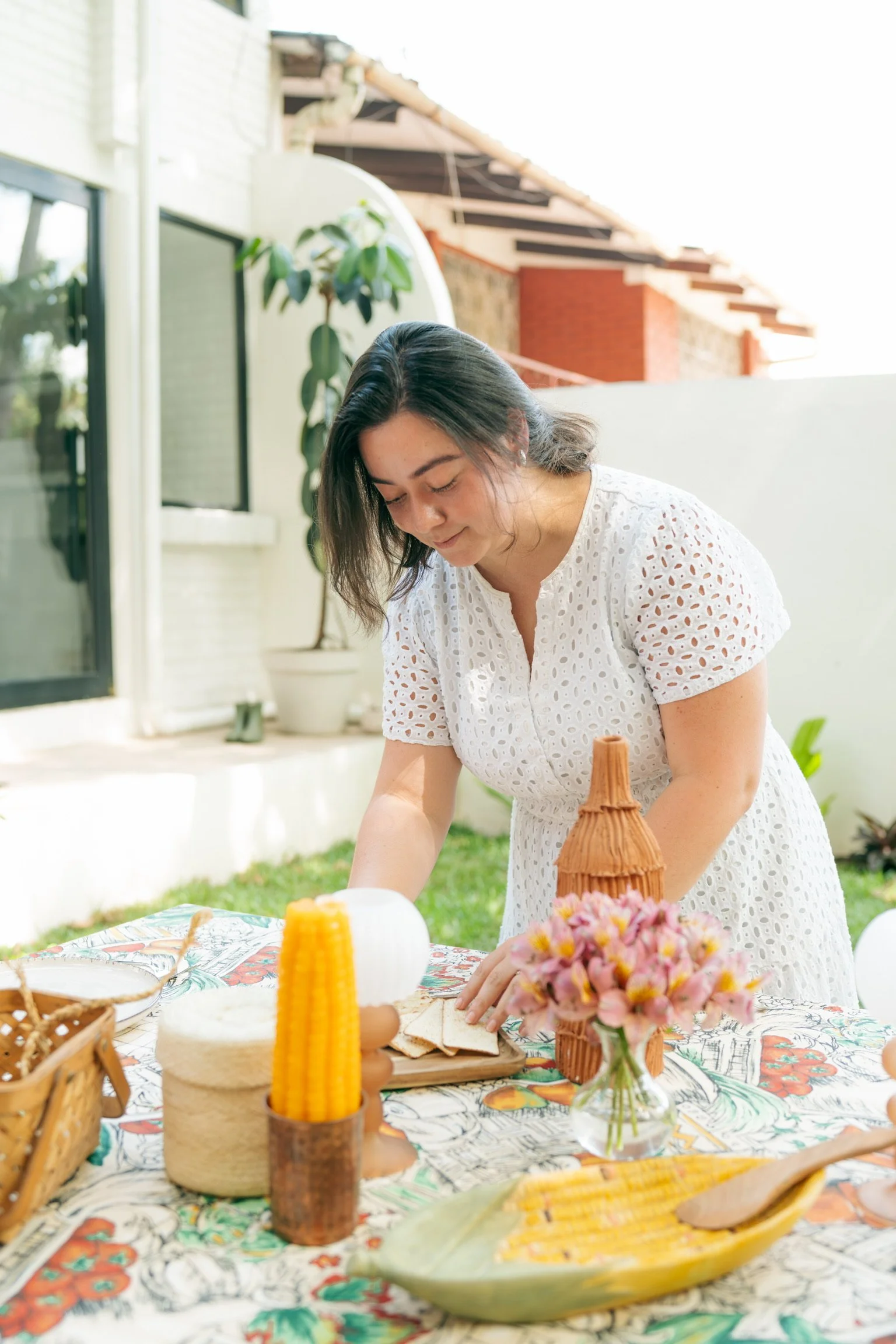 A woman in a white eyelet dress arranging a table outside with colorful tablecloth, flowers, and food items.