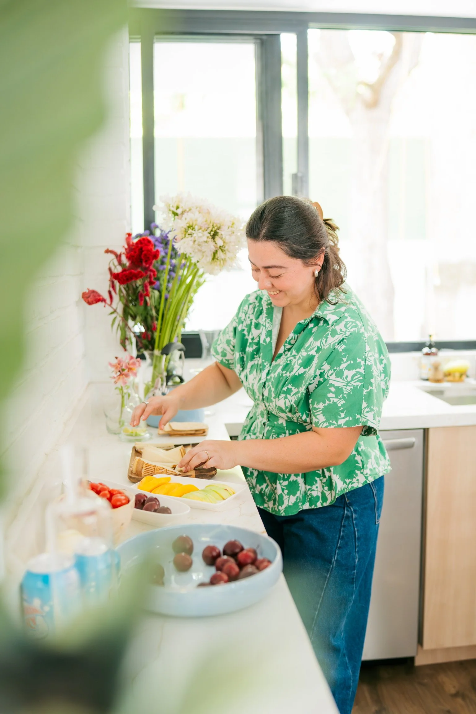 Woman in a green patterned shirt preparing fruit on a kitchen countertop, with a vase of colorful flowers behind her and a window in the background.