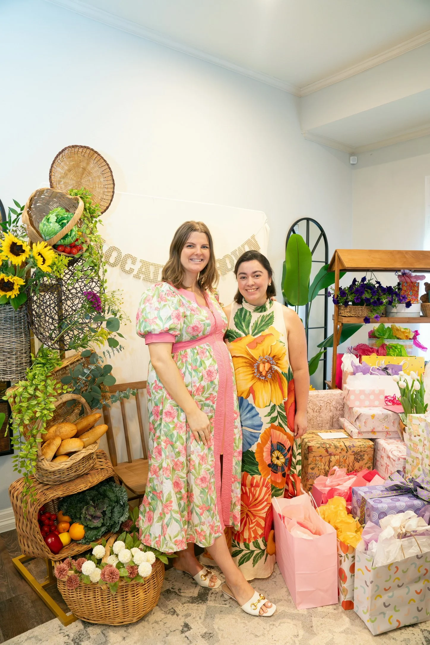 Two women standing among colorful flowers, gift bags, and decorative items at a celebration or event.