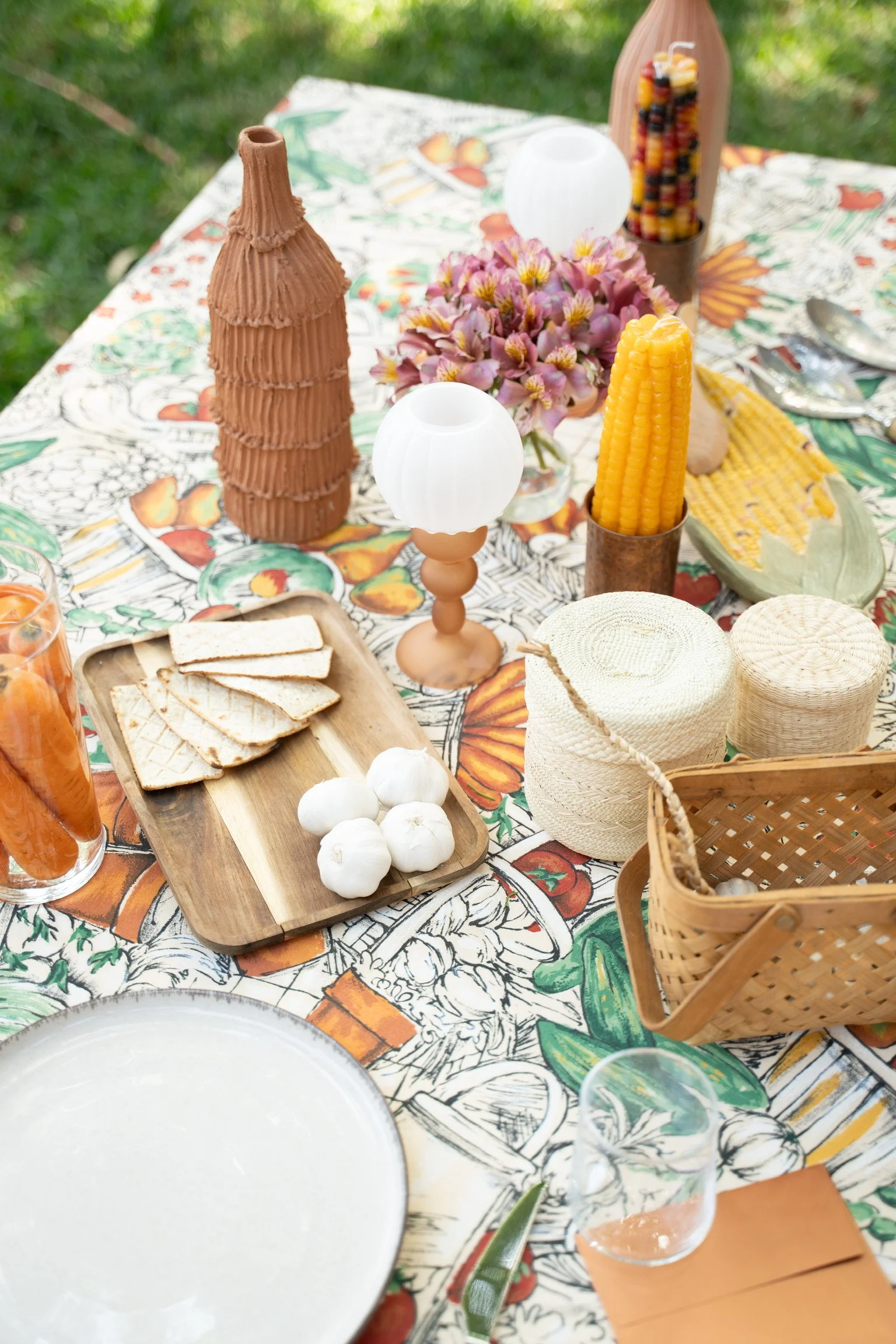 Outdoor table set with a colorful floral tablecloth, decorated with a brown vase resembling a tiered cone, white spherical lamps, a cut honey crisp corn, a bunch of purple flowers, a wooden tray with sliced bread and garlic, glasses with beverages, and wicker baskets.