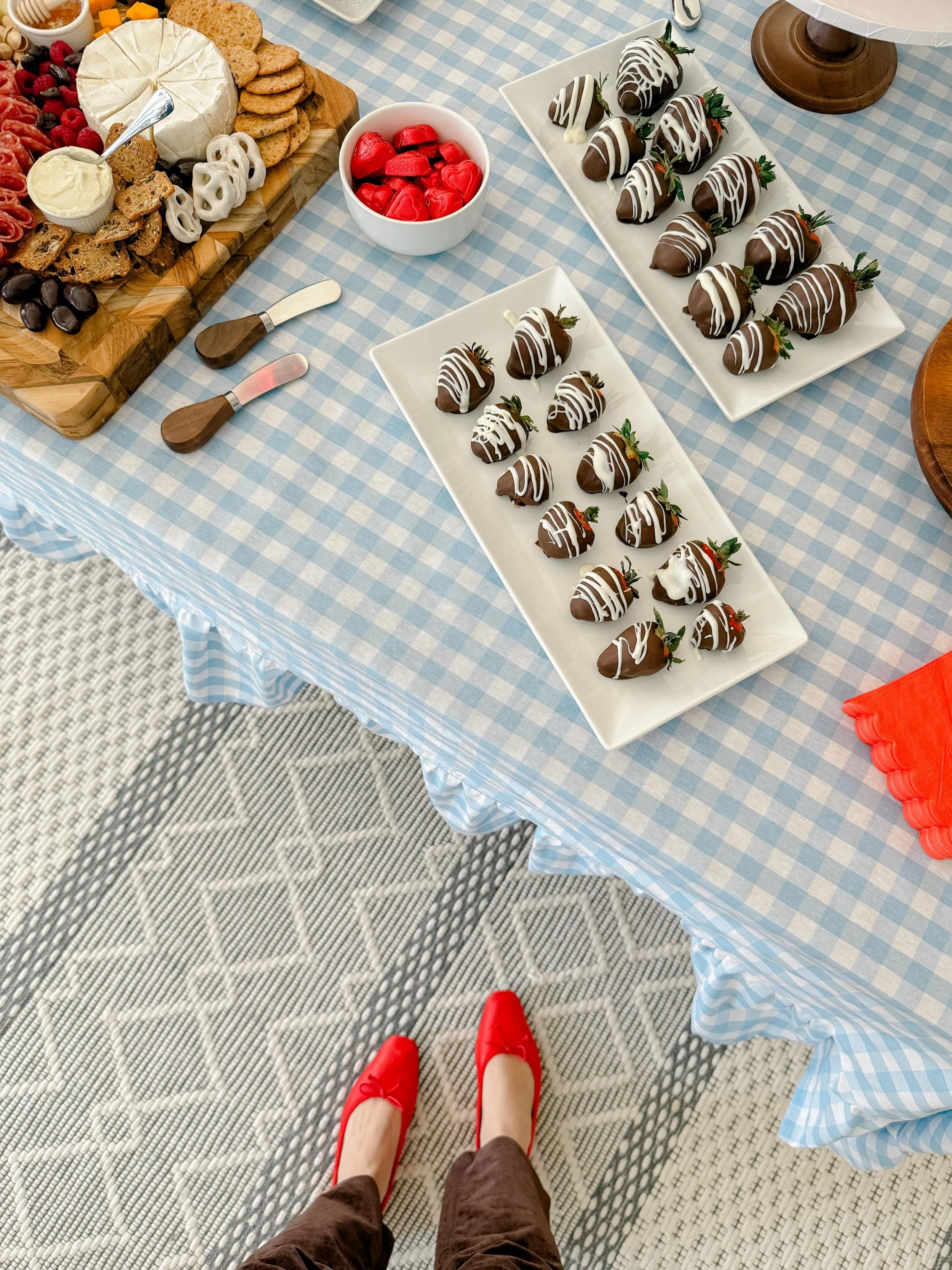 A table with a blue and white checkered tablecloth holds a cheese and charcuterie platter, a bowl of red candies, and two white rectangular plates with chocolate-covered strawberries topped with white chocolate drizzle and green stems. The picture is taken from the perspective of a person wearing red shoes and brown pants, standing on a gray and white patterned rug.