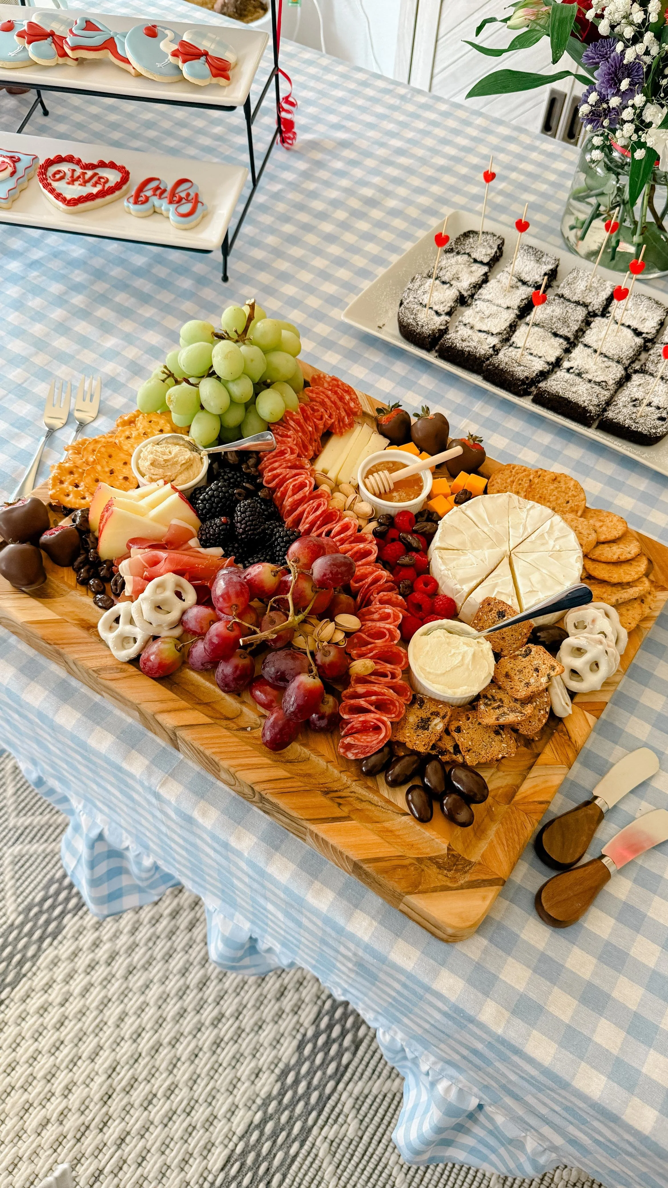 A cheese and fruit platter with grapes, crackers, sliced meats, cheeses, and berries on a wooden board, with small desserts and cookies on a table covered with a blue and white checkered tablecloth.
