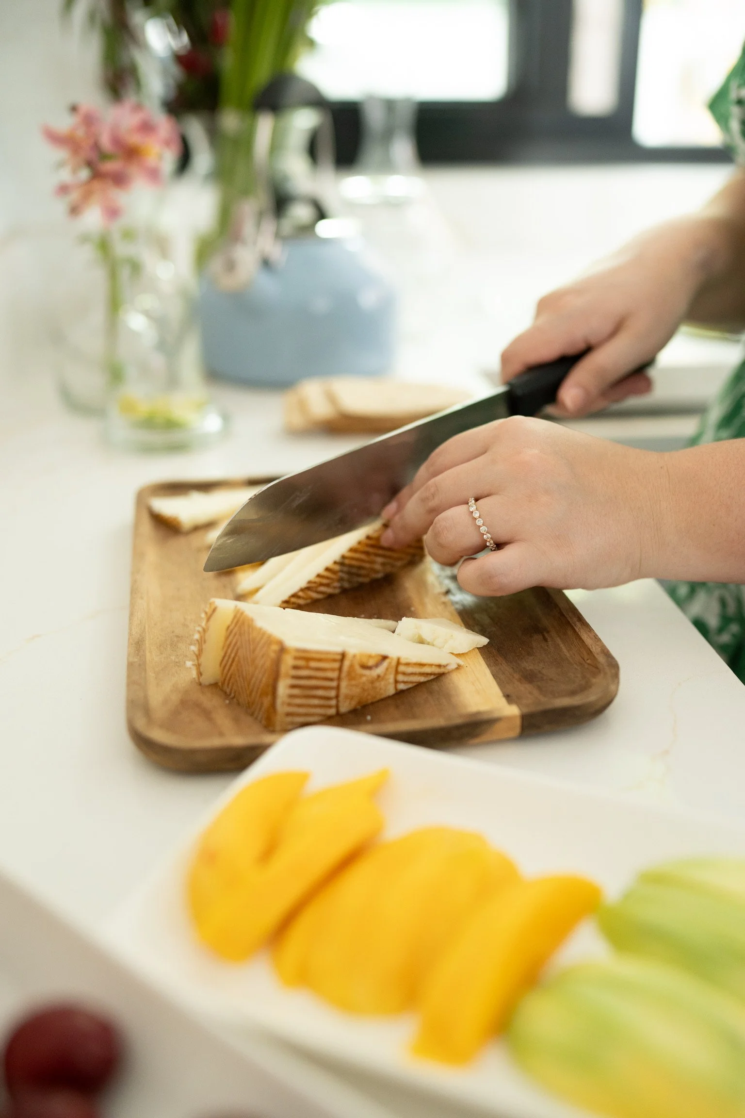 Person slicing cheese on a wooden cutting board in a bright kitchen, with flowers in a vase and a window in the background, and a fruit platter in the foreground.