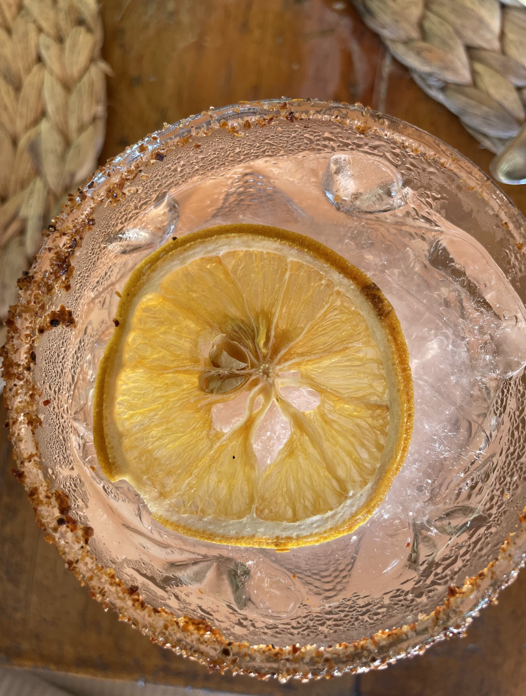 A top-down view of a glass of water with ice and a slice of lemon, with a salt or chili rim on the glass, placed on a wooden surface.