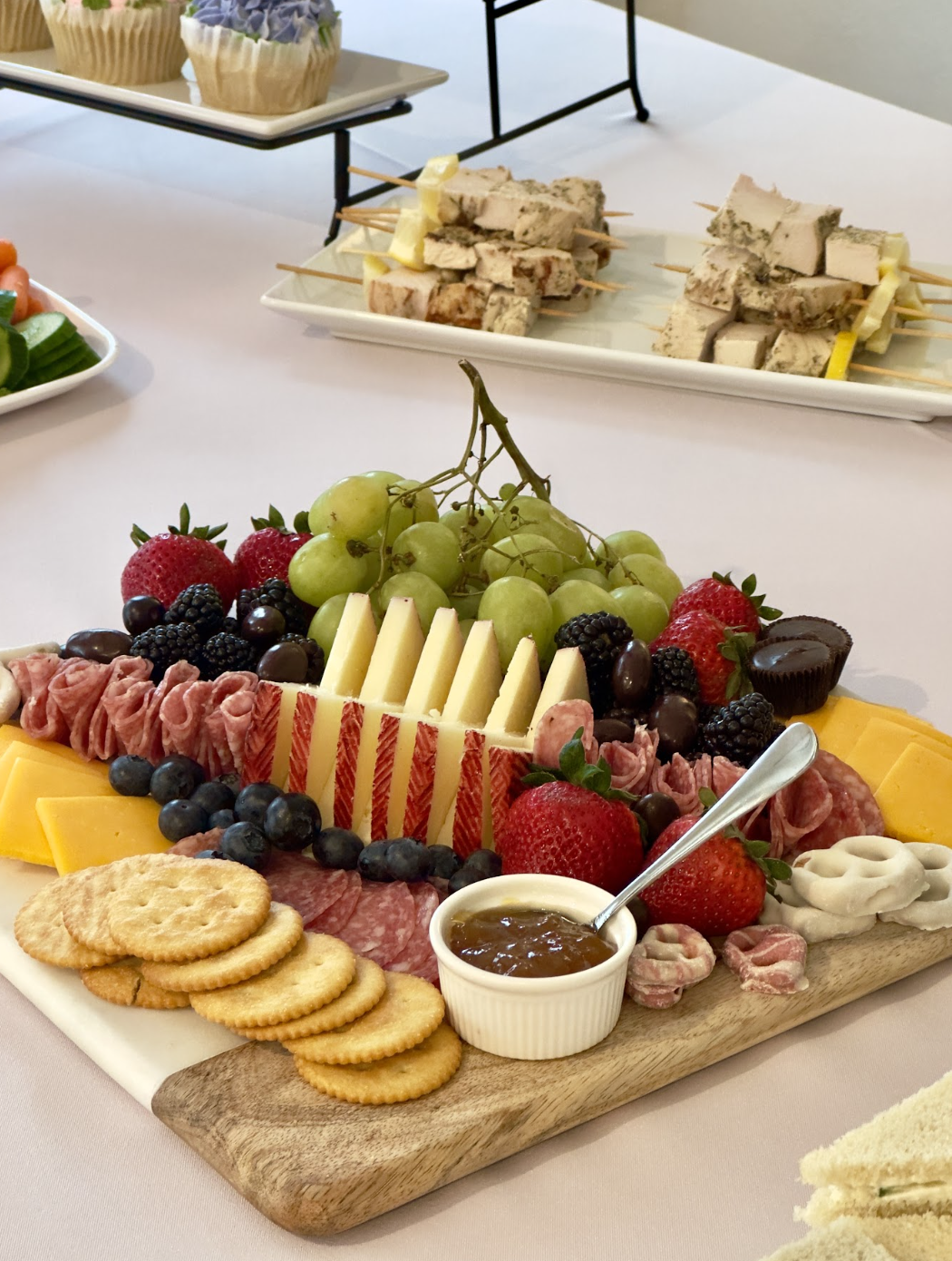 Antipasto platter with assorted cheeses, cured meats, fresh strawberries, grapes, blueberries, blackberries, black olives, and a small bowl of jam. Accompanying crackers and slices of cheddar cheese are also on the wooden board.