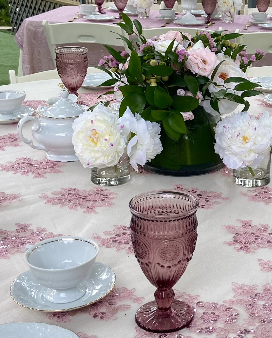 A pink floral tablecloth with a centerpiece of pink and white flowers, green leaves, and berries. The table is set with white china, a white teapot, pink tinted glasses, and pink napkins, arranged for a formal event or tea party.
