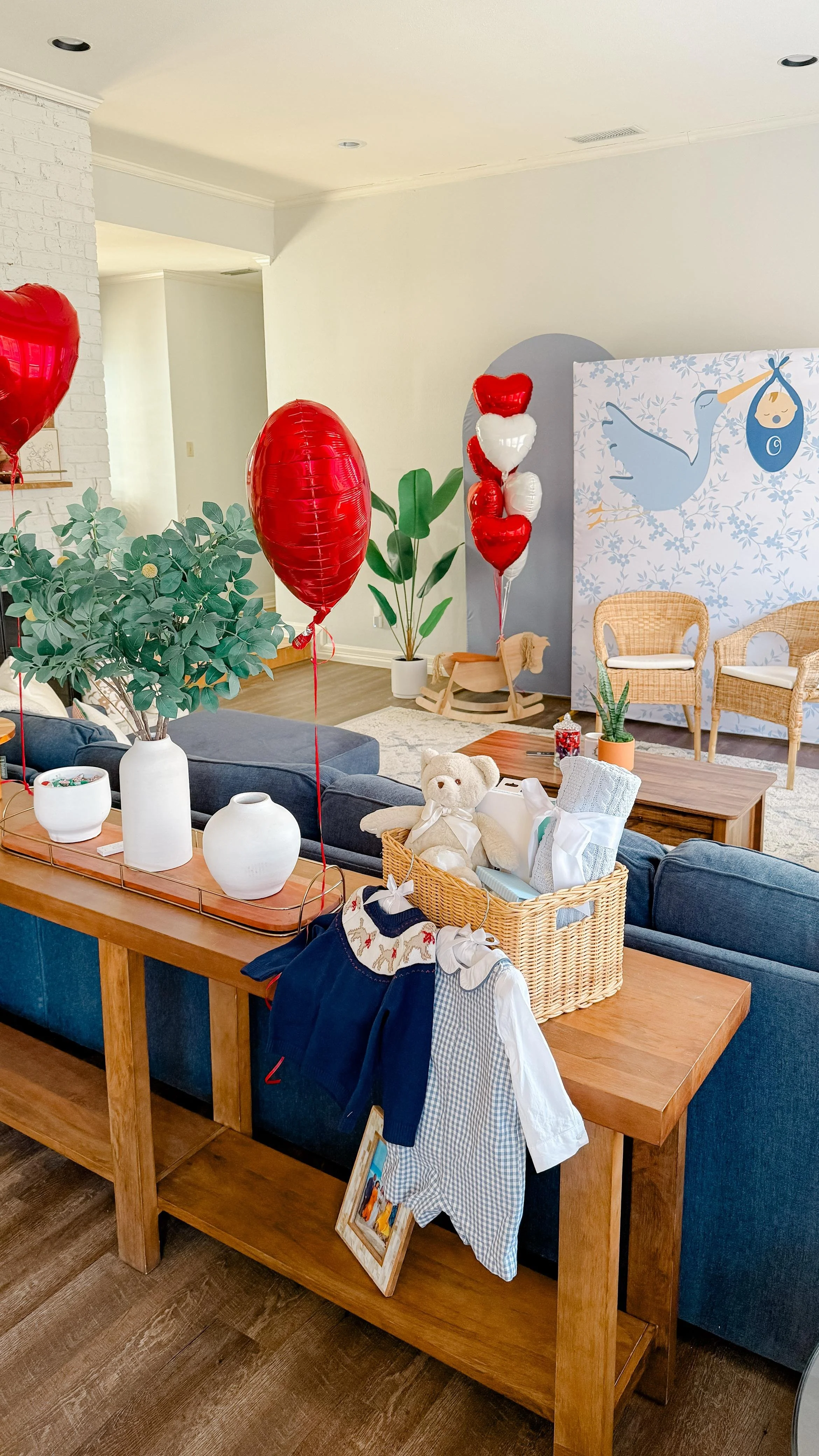 Living room decorated for a celebration with red and white heart-shaped balloons, potted plants, a teddy bear, gift boxes, and playful baby clothes hanging on a table.