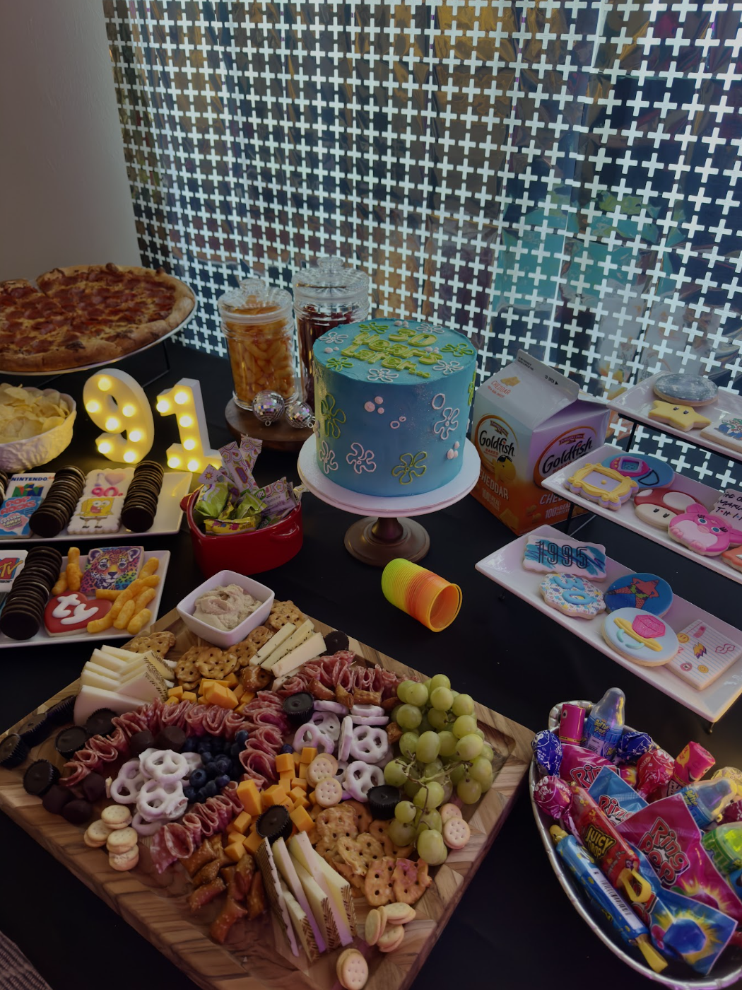 A birthday party setup with a table featuring a blue floral cake, pizza, a charcuterie board, various candies, and party decorations such as a large illuminated "91" sign, with a backdrop of patterned curtains.