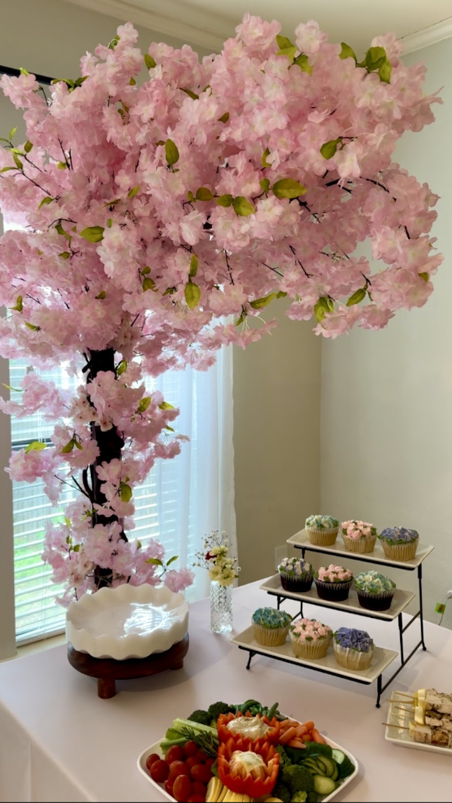 Decorative pink flowering tree placed next to a table with assorted cupcakes, a vegetable platter, and a small tray of finger foods, with a window and white curtains in the background.