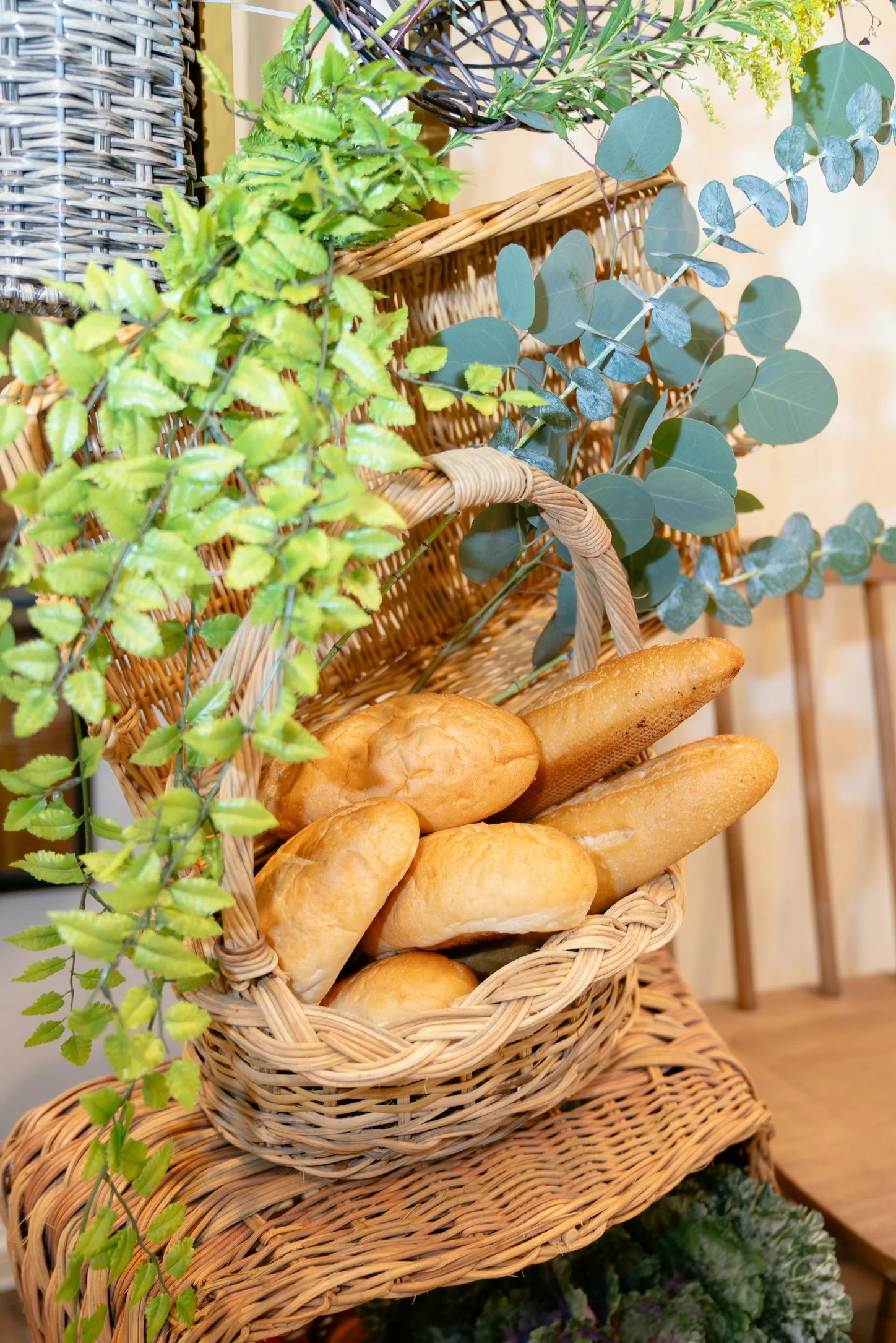 A close-up of a wicker bread display stand with multiple shelves holding assorted bread rolls and baguettes, decorated with green leafy plants.