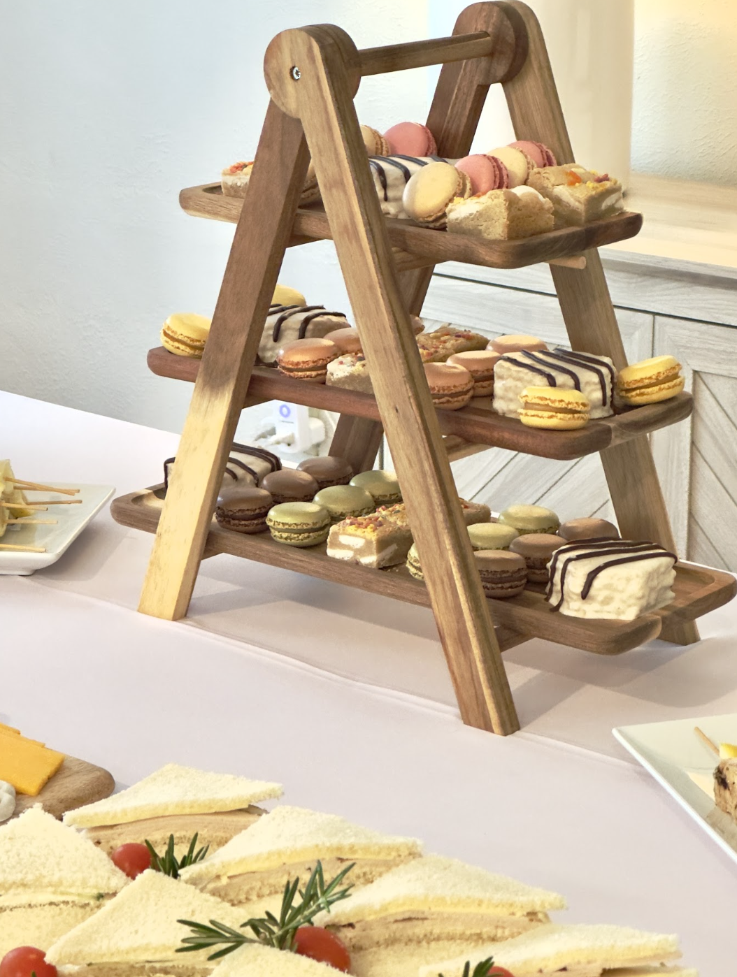 A wooden three-tiered dessert stand filled with assorted macarons and small slices of cake on a white table.