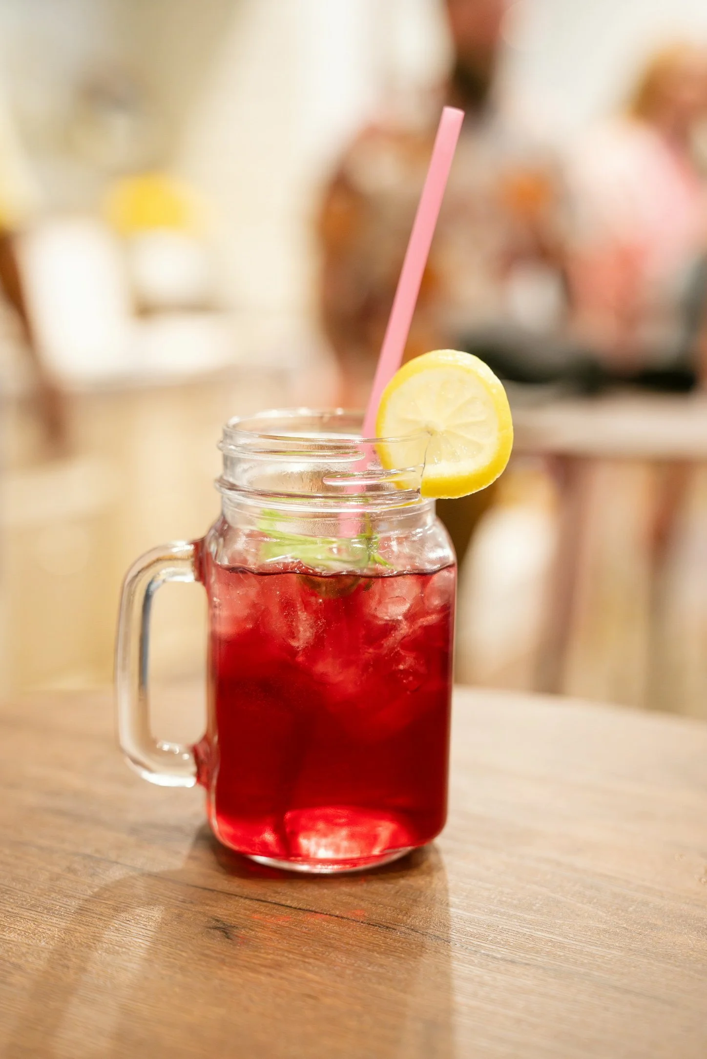 Red beverage in a mason jar glass with lemon slice garnish and pink straw, on a wooden table.