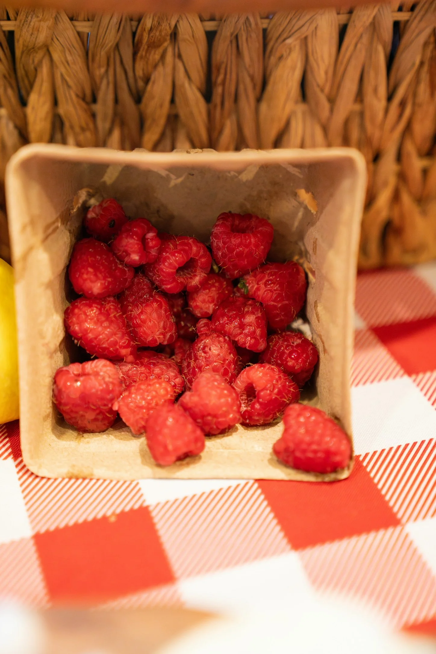 Fresh red raspberries inside a small square wooden container on a red and white checkered cloth.