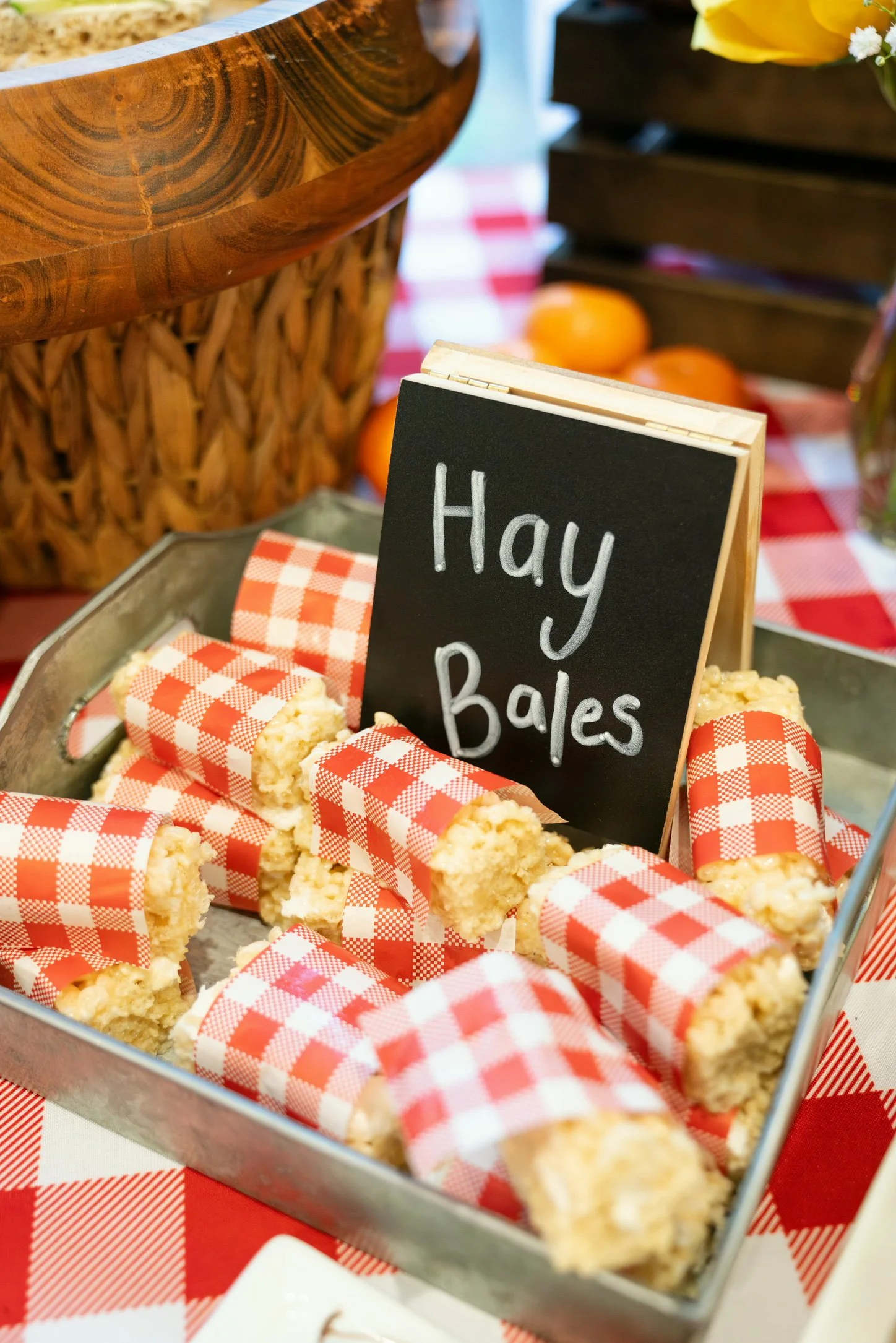 A metal tray filled with cherry baked bars wrapped in red and white checkered paper, with a blackboard sign that says "Hay Bales" behind them. In the background, there are decorative elements including a woven basket, a wooden crate, oranges, and flo