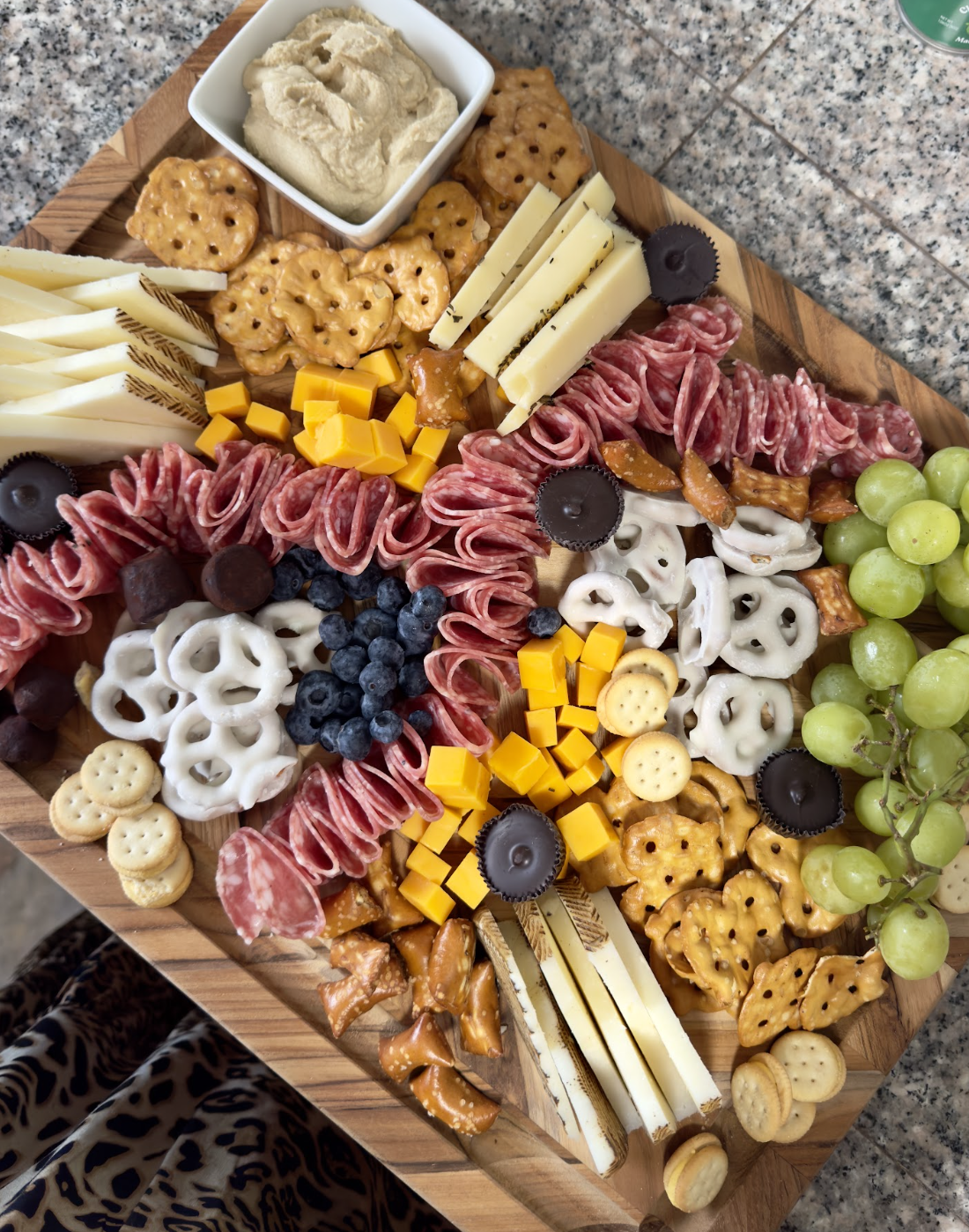 A wooden cheese and charcuterie platter featuring sliced cheeses, cured meats, grapes, blueberries, crackers, cheese cubes, and cheese slices, accompanied by a bowl of hummus and small crackers.