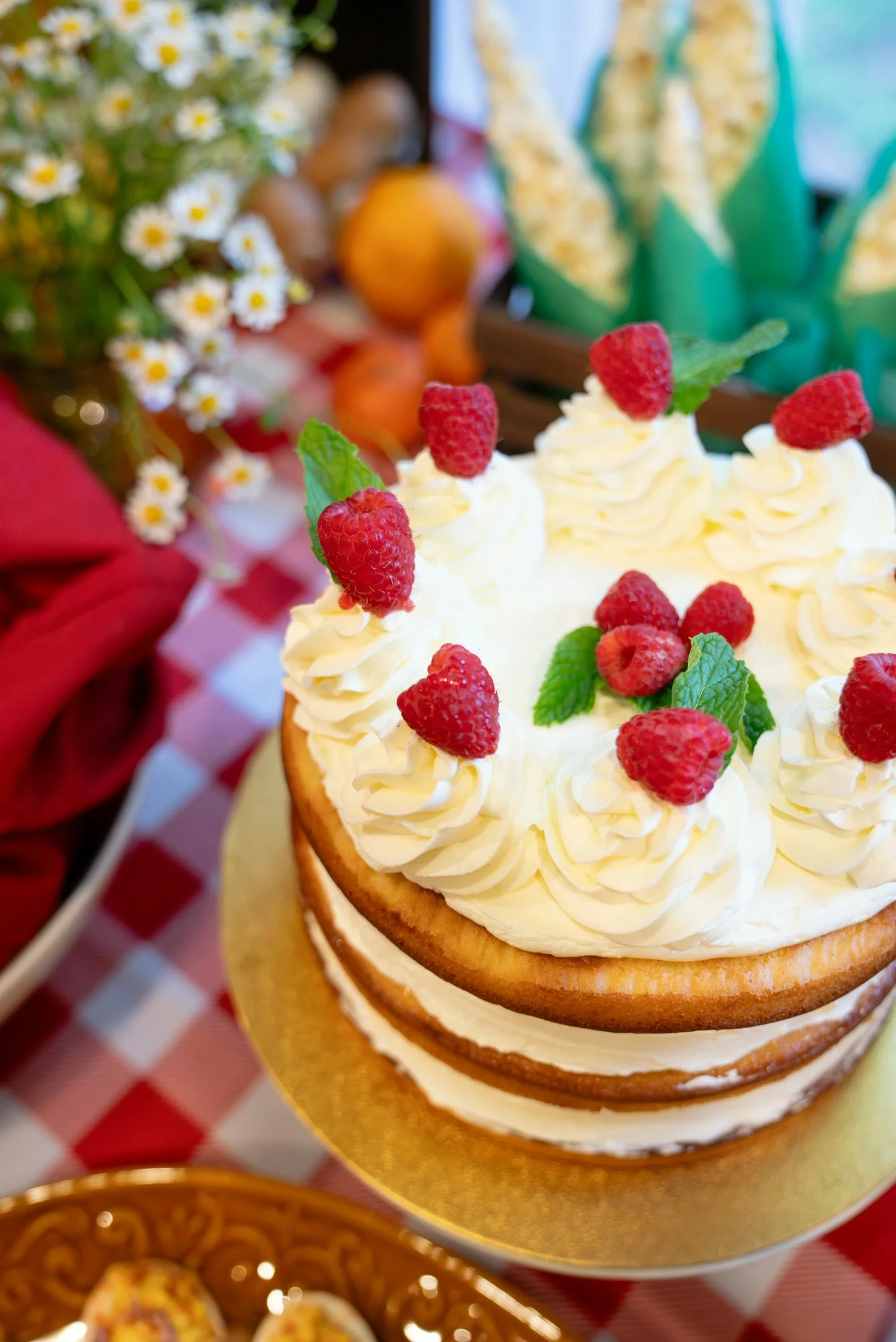 A cake decorated with raspberries, mint leaves, and cream on top, with a blurred background of flowers and other items.