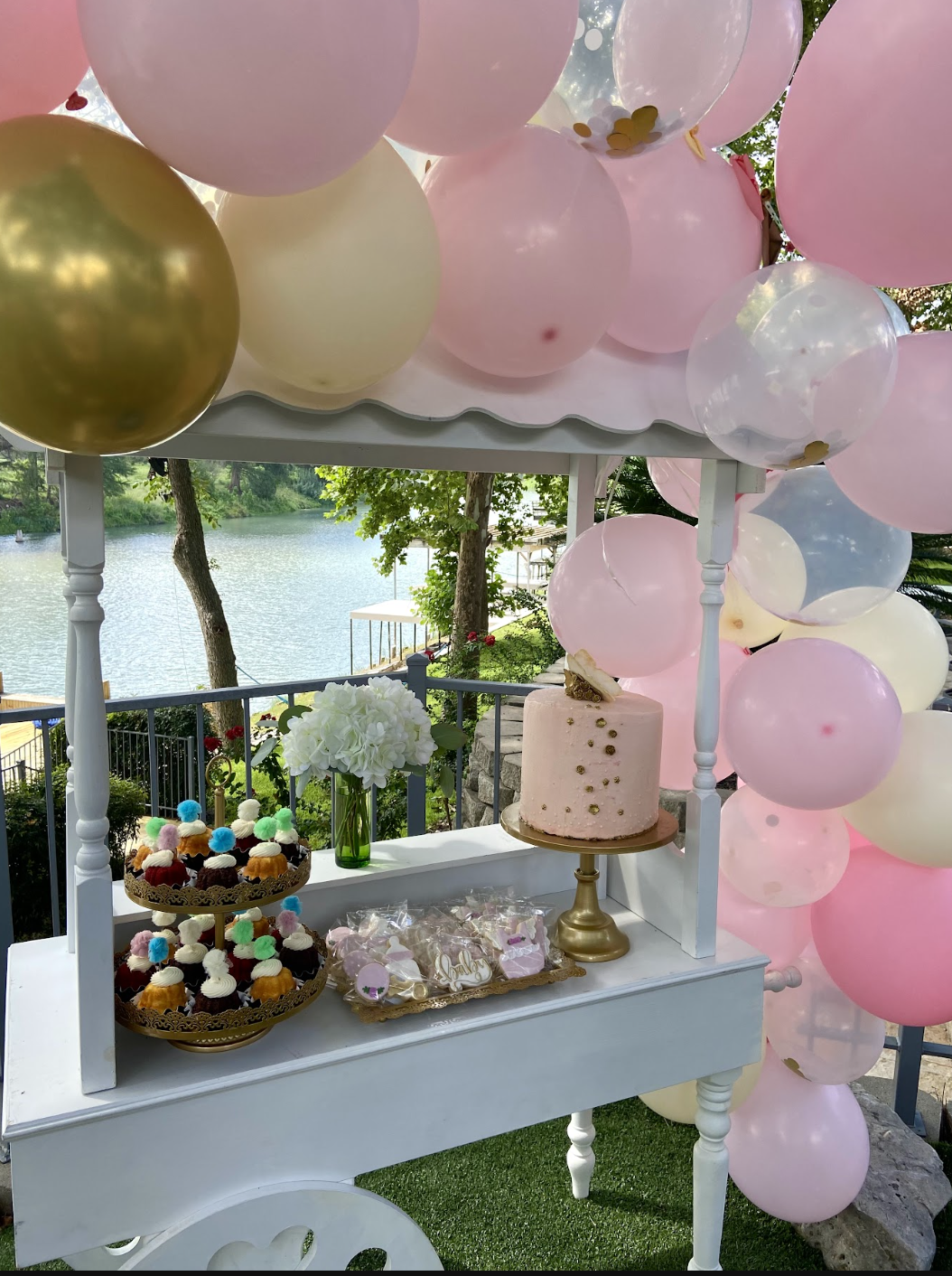 Decorated cart with pink, white, and gold balloons, a pink cake on a golden stand, cupcakes with colorful toppers, a green vase with white flowers, and a waterfront view with trees and a railing.