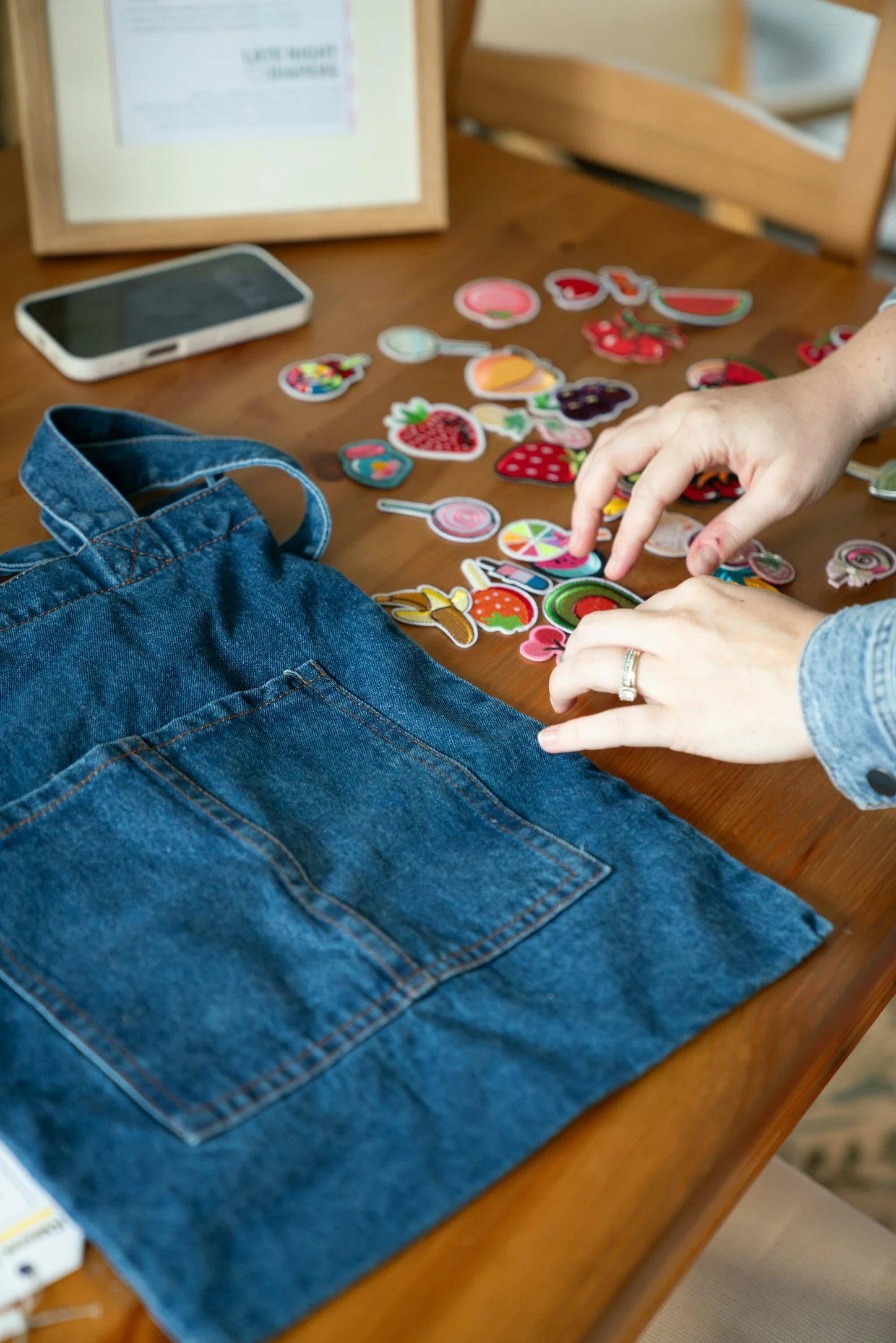 A person with light skin and a wedding ring is placing or picking up embroidered patches shaped like various fruits and drinks on a wooden table. There is a blue denim jacket in the foreground, a white smartphone to the left, and a blurred framed doc