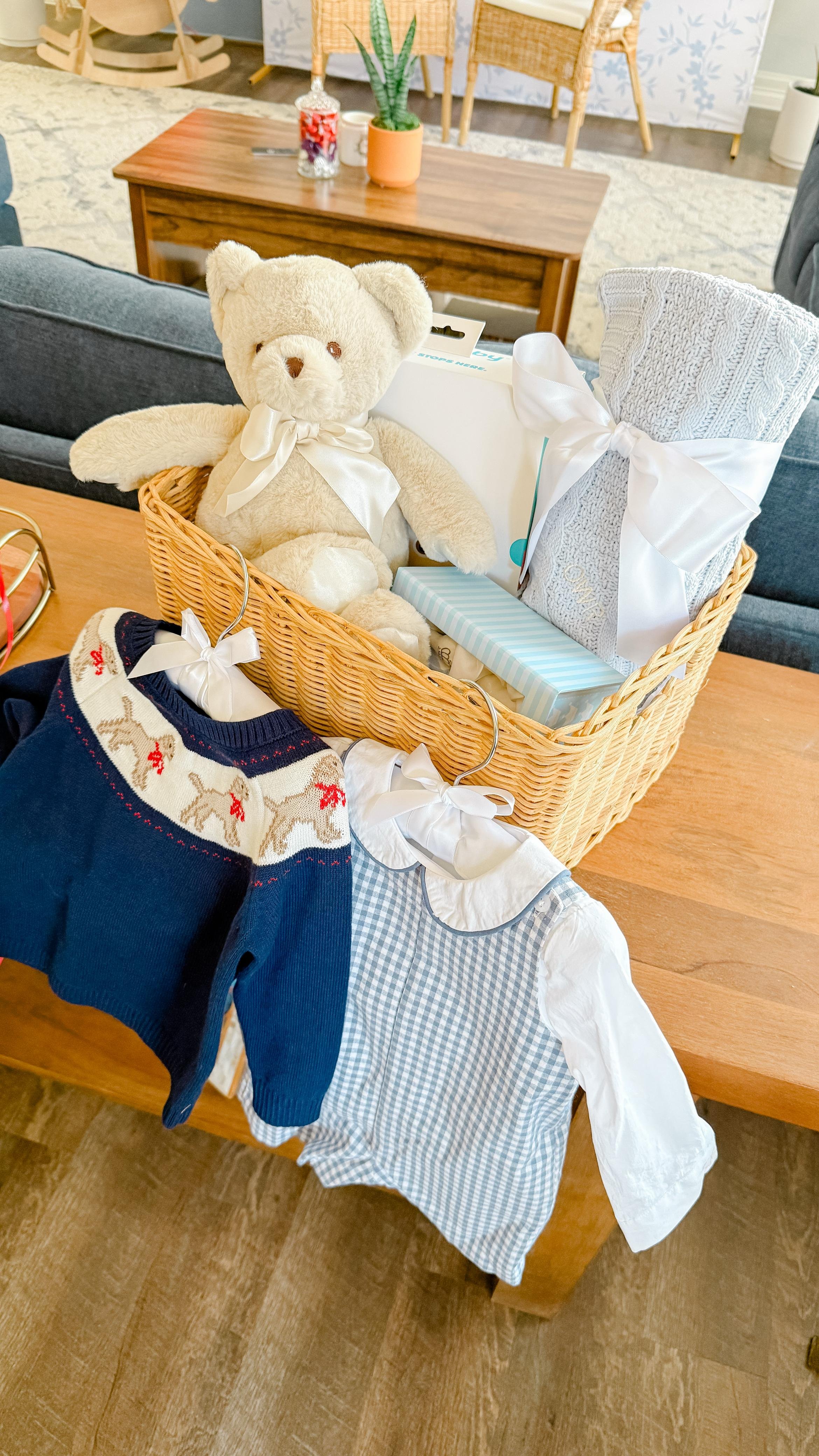 A wicker basket on a wooden table contains a teddy bear, a rolled-up white towel tied with a ribbon, and other wrapped gifts. In front of the basket, on the table, are Christmas socks, one with reindeer pattern and the other plain white.