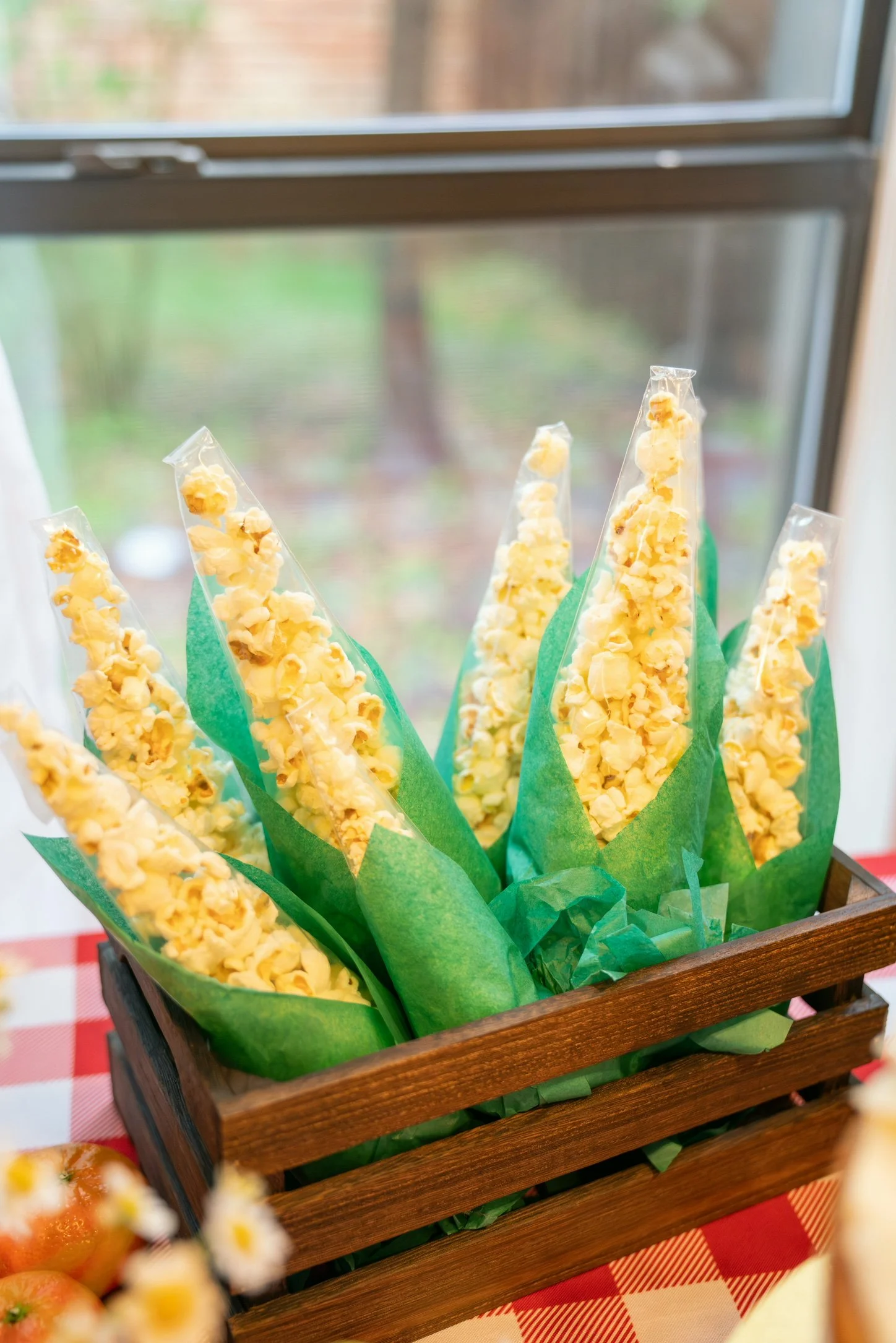 Yellow and white popcorn in clear plastic bags wrapped with green paper, displayed in a wooden basket on a red and white checkered tablecloth.