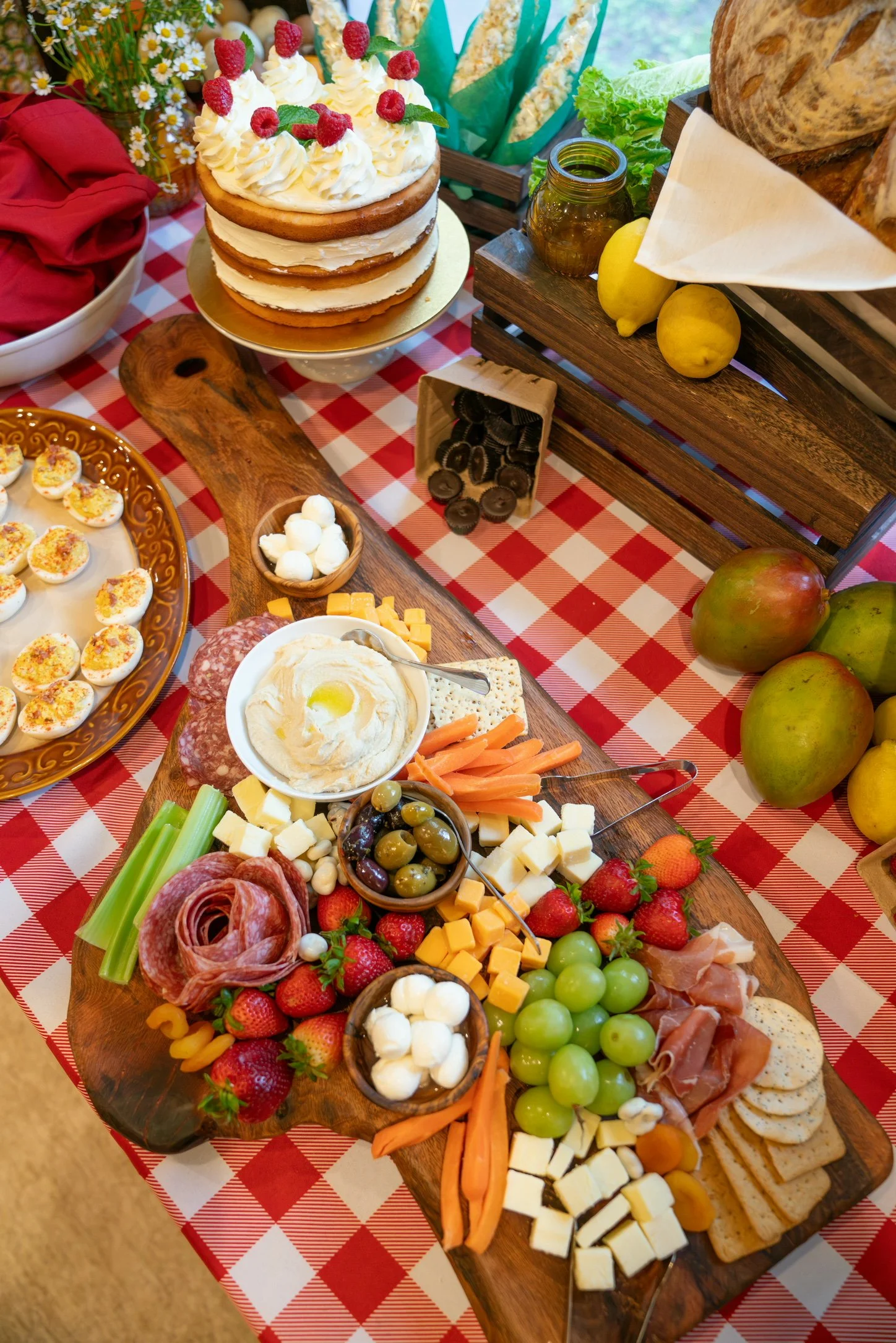 A spread of food items including a layered cake decorated with raspberries and mint, deviled eggs with paprika, cheese cubes, olives, sliced meats, fresh strawberries, green grapes, carrots, celery, cheese, bread, and a bowl of cow's milk cheese, all