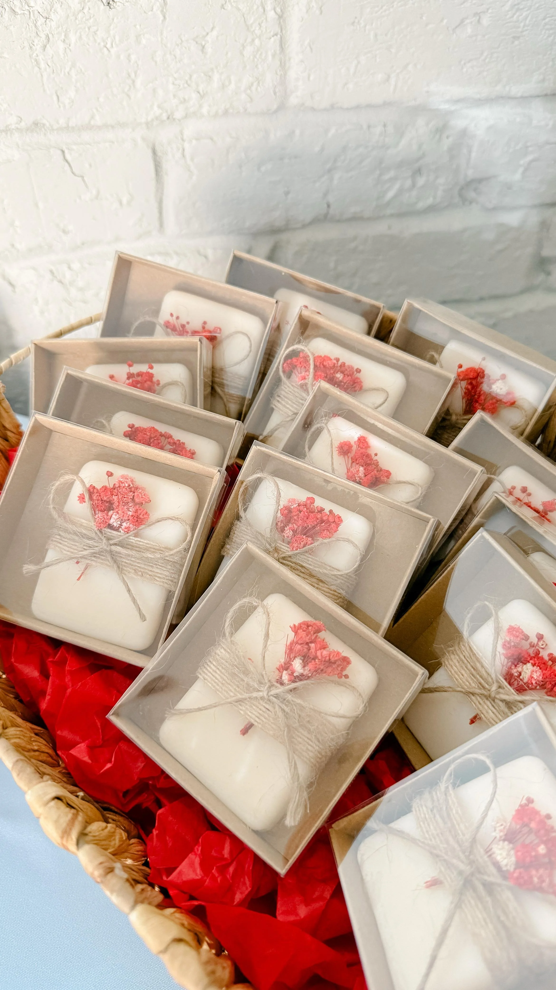 A basket filled with wrapped candles decorated with pink flowers and twine, placed on red tissue paper.