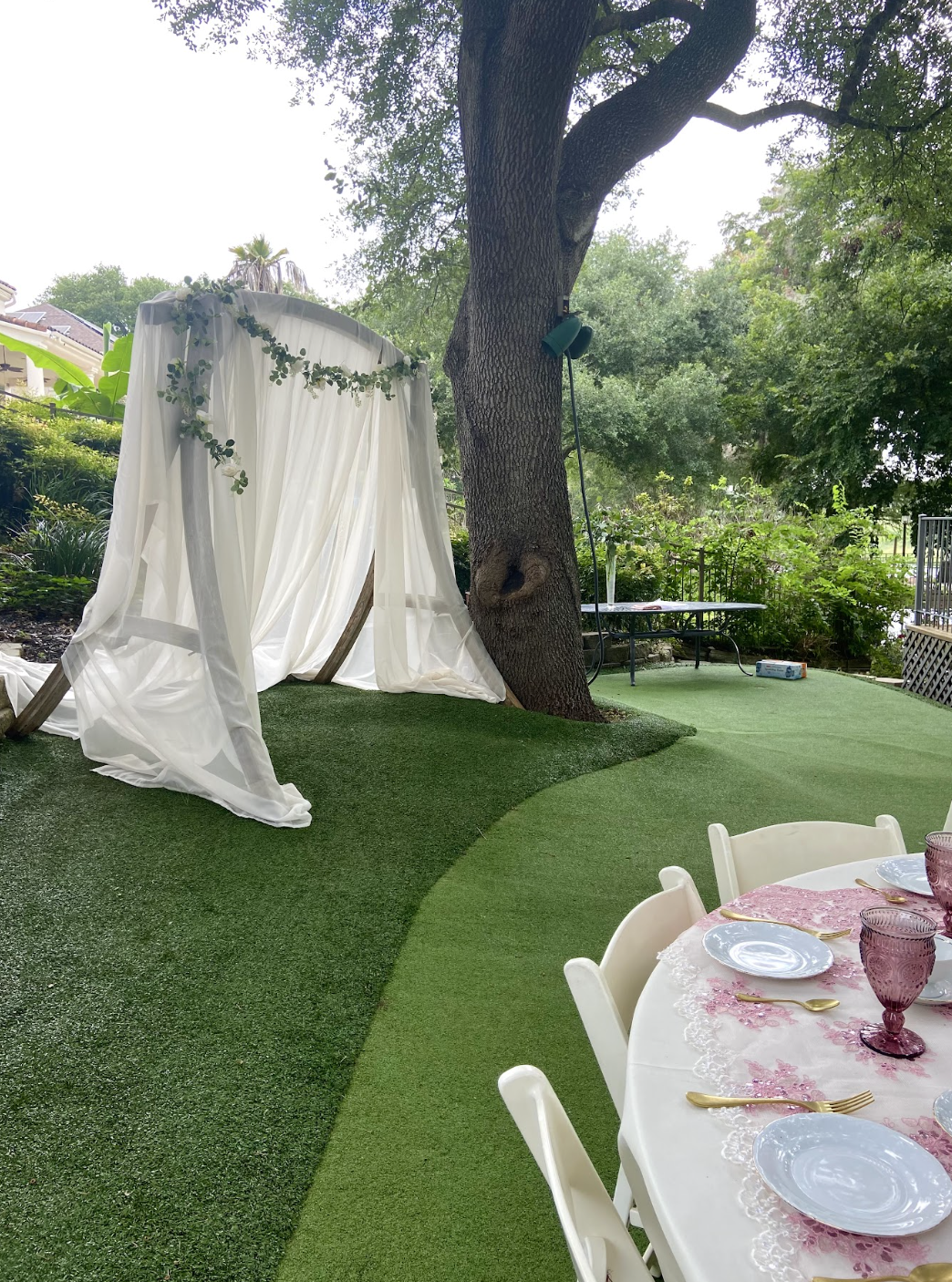 Outdoor setting with a white canopy decorated with greenery, a large tree, and a dining table set with pink and purple glassware and white plates, indicating a celebration or event.