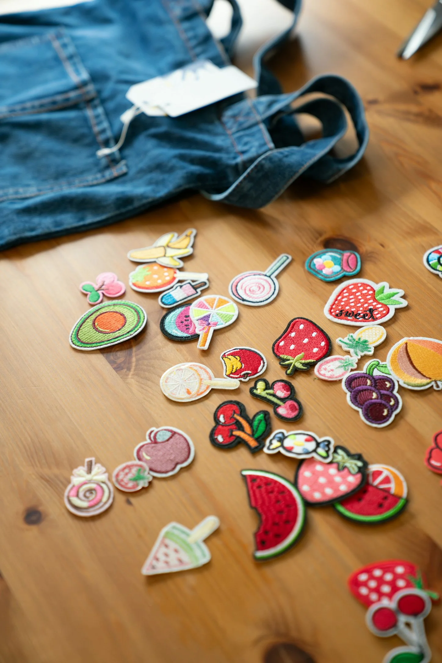 Colorful embroidered patches of various fruits and sweets scattered on a wooden surface, with a denim bag nearby.