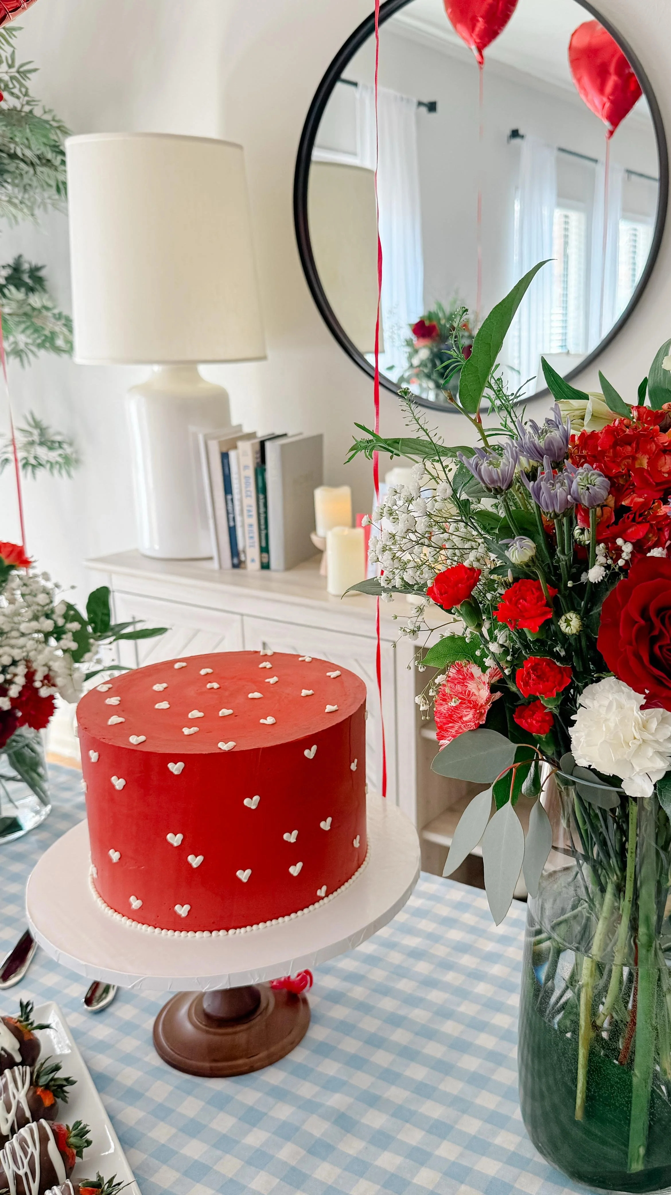A red frosted cake decorated with small white hearts on a cake stand, surrounded by a large bouquet of red, white, and purple flowers in a green vase, with balloons in the background.