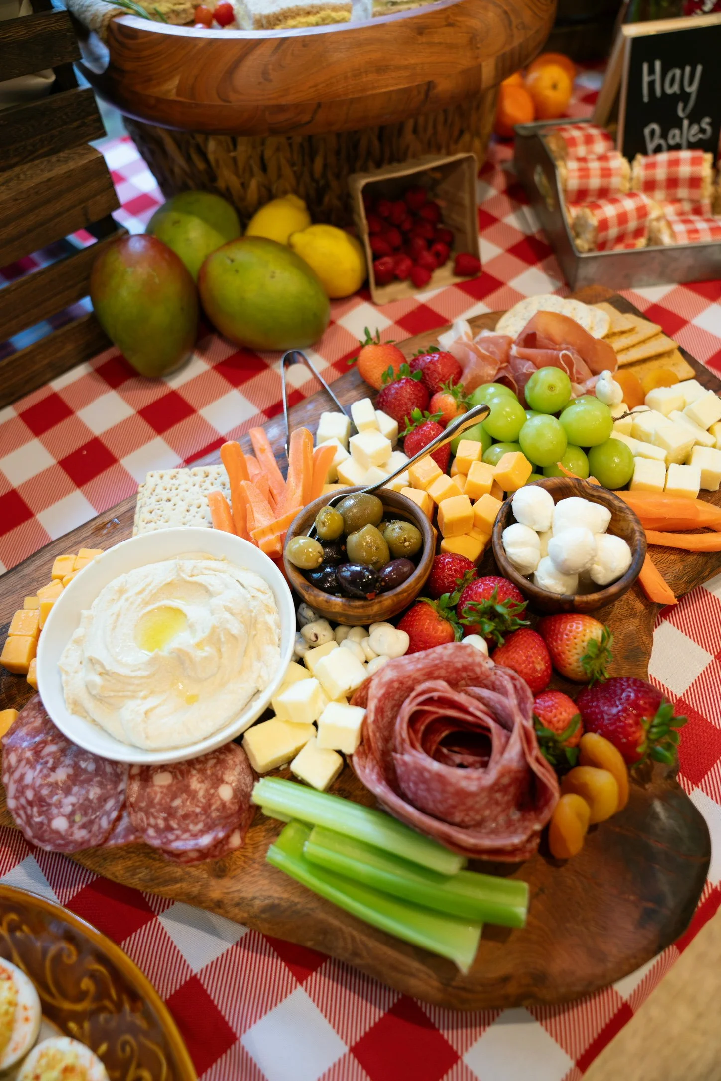 A cheese, fruit, and snack platter on a wooden board with strawberries, grapes, cheese cubes, sliced cured meats, carrot sticks, celery, mozzarella balls, and chip dip, with a cheese and cracker container in the background.