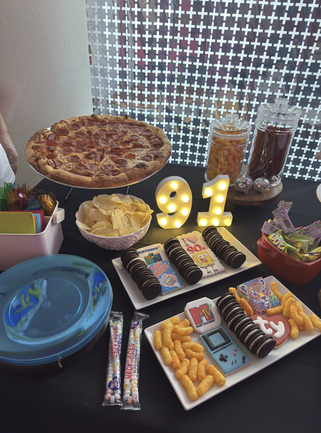 Table set with pizza, potato chips, cookies, popcorn, candies, and soda bottles, decorated with a 91 birthday number sign for a celebration.
