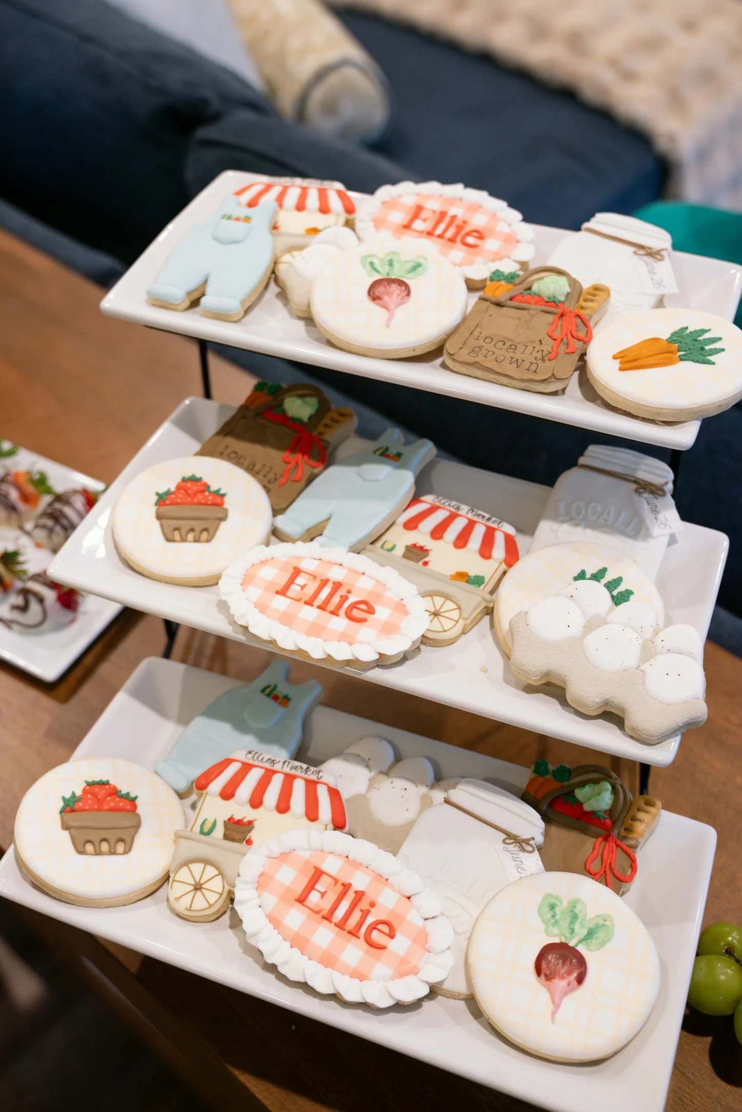 Assorted decorated cookies on white trays, including cookies with vegetables, a shopping bag, and the name 'Ellie' in red on a gingham pattern.