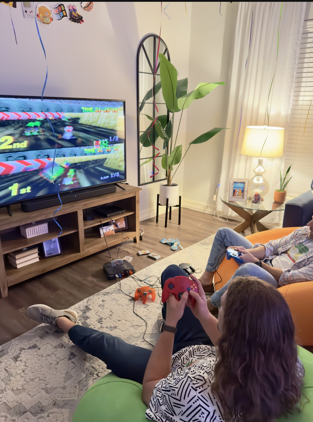 Two children playing a Nintendo 64 video game in a living room. One is holding a red controller, and the other is holding a blue controller. The TV screen shows a racing game with a first and second place. The room is decorated with balloons, with a 