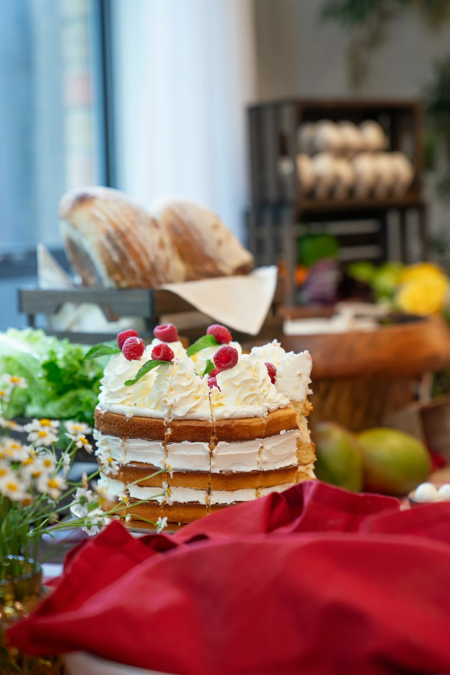 Sweet layered cake topped with whipped cream and raspberries, surrounded by flowers and red cloth with bread in the background.