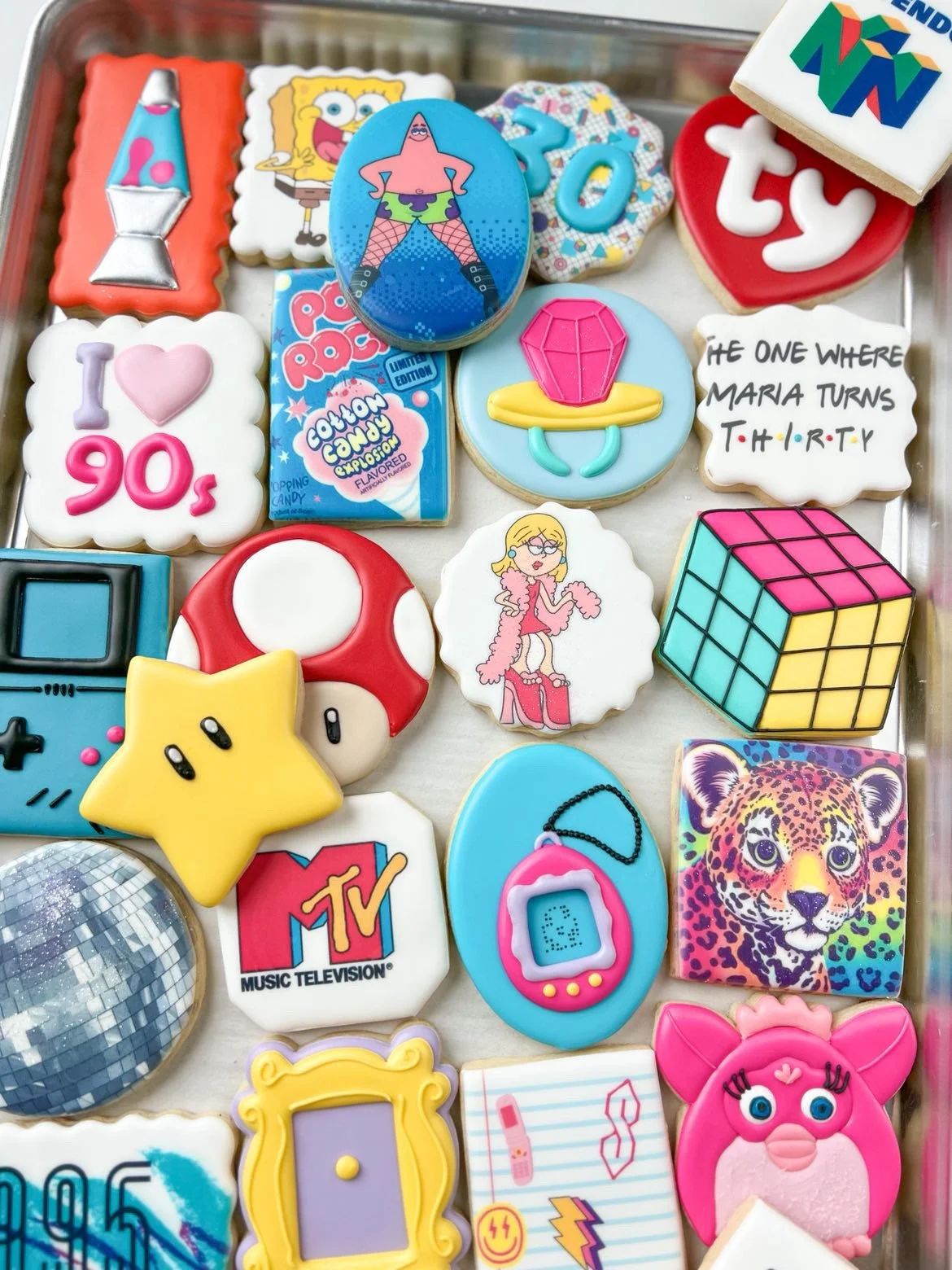 Collection of decorated cookies with various pop culture and nostalgic themes, including characters from SpongeBob SquarePants, a Rubik's Cube, a Game Boy, a Tamagotchi, a leopard, a star, a heart, and text-based cookies celebrating the 1990s.