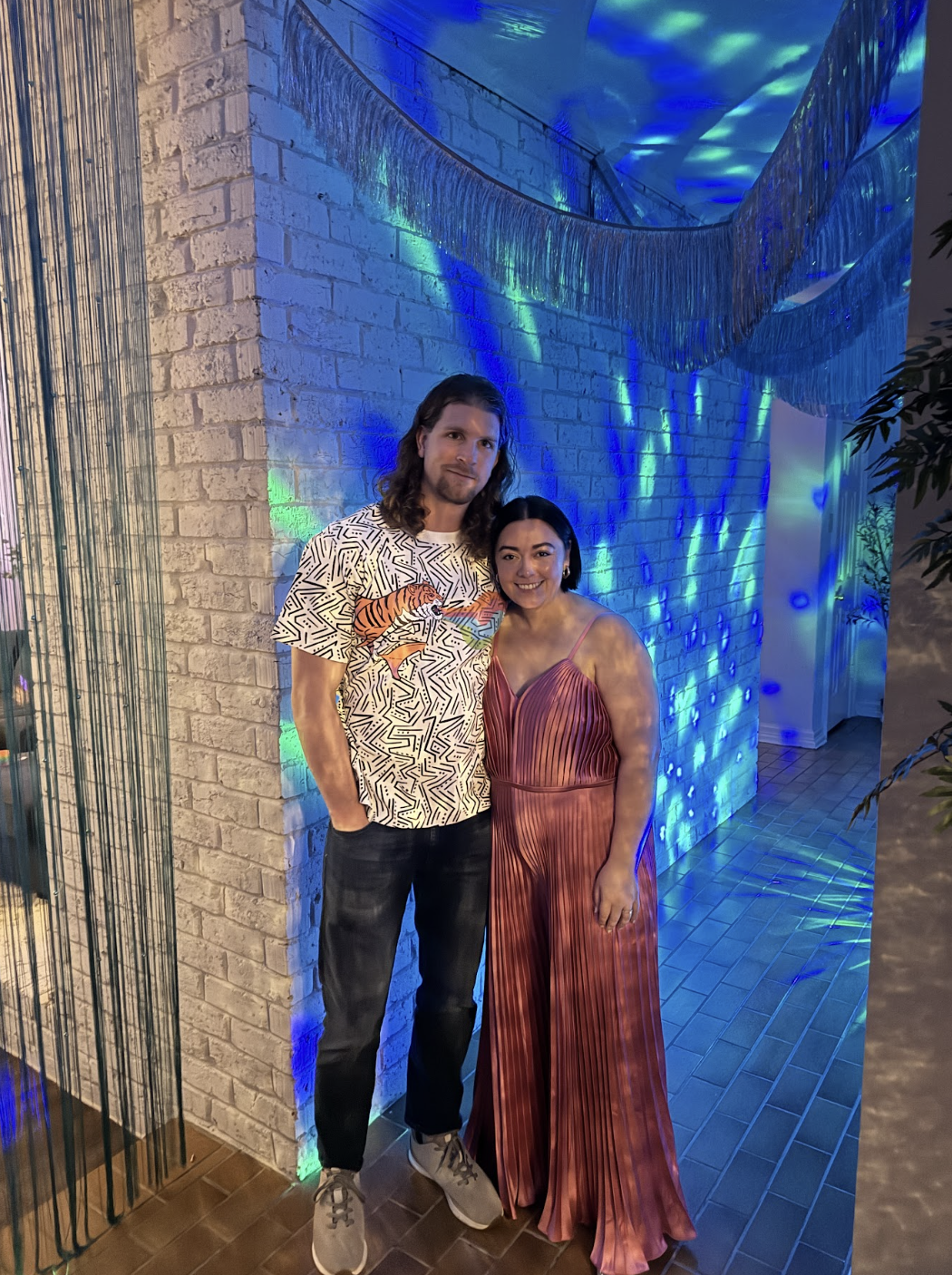A man and woman are standing together indoors near a brick wall with colorful blue and green lighting. The man has long hair and is wearing a patterned T-shirt with a tiger and other designs, black pants, and gray sneakers. The woman has short black 