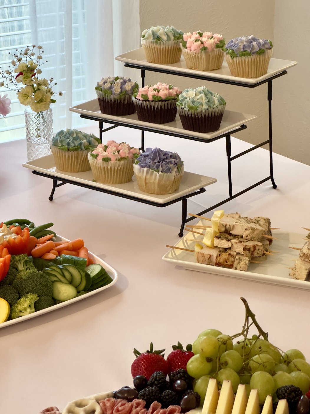 Dessert display with assorted cupcakes in pastel colors on a tiered stand, fresh fruit, vegetable platter, and skewered appetizers on a white tablecloth.